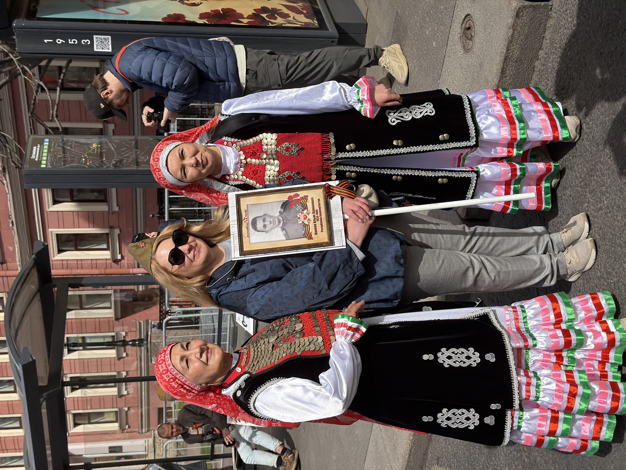The image shows three women in traditional folk costumes standing on a street. The two women on either side of the image are wearing similar traditional outfits, with black vests adorned with decorative metal elements over white blouses and layered skirts with pink, white, and green stripes. The woman in the middle is wearing a blue jacket, gray pants, and a military-style hat. She's holding a framed photograph with a ribbon attached.
A man is taking a picture of a building behind the women. The background contains buildings typical of Saint Petersburg, Russia, with their architectural style. There is also a bus stop sign in the scene. The environment suggests an urban setting.