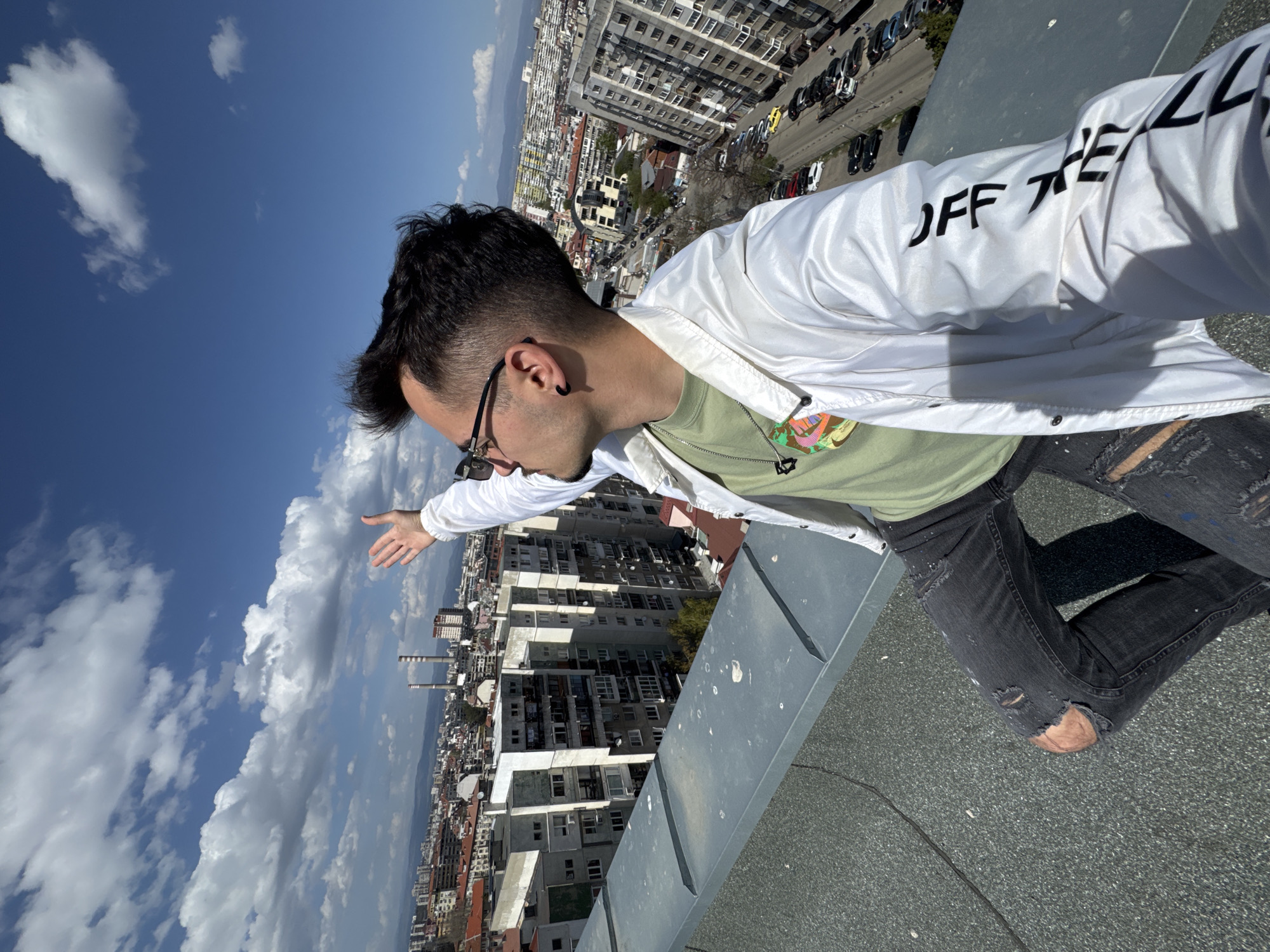 The image features a young man leaning precariously over the edge of a building's rooftop, looking out over a bustling city on a clear, sunny day. He is positioned on his right side, with his left arm extended outwards, seemingly gesturing towards the panorama below.
The man is dressed in a white jacket with the text "OFF THE LL" prominently displayed in black capital letters on his left sleeve. Beneath the jacket, he wears a light green t-shirt and dark, distressed jeans with visible rips at the knees. He has short, dark hair, wears dark-rimmed glasses, a black ear gauge, and a black pendant necklace.
The setting is a vibrant urban environment, consistent with Sofia, Bulgaria. From the high vantage point of the rooftop, a dense cityscape unfolds, filled with numerous multi-story buildings, roads, and parked cars. In the distance, mountains line the horizon, and two tall industrial chimneys are visible. The sky is a brilliant blue, adorned with scattered white, cumulus clouds. The rooftop itself is a flat, dark grey, textured surface, with a distinct grey metal ledge that the man is leaning against. The bright sunlight suggests it is daytime. The overall scene conveys a sense of adventure, freedom, and an appreciation for the extensive city view.