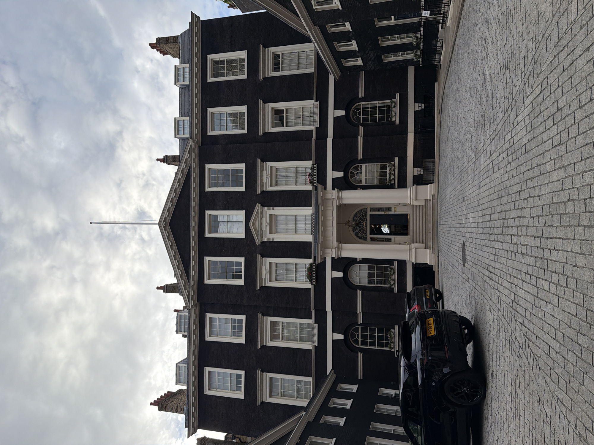 The image depicts the facade of a grand, dark brick building in London, United Kingdom, likely a Georgian-era townhouse or a formal institutional building. The building features symmetrical architecture with numerous large, multi-paned windows, white trim, and a prominent central entrance with columns and a pediment. Chimneys are visible on the roofline, and dormer windows are interspersed. A flagpole stands at the apex of the central gable.
In the foreground, a cobbled courtyard is visible, with two black cars parked on the left. The car closest to the viewer is a Range Rover, with a yellow license plate. The sky is overcast with clouds. The overall impression is one of stately elegance and historical architecture in an urban setting.
