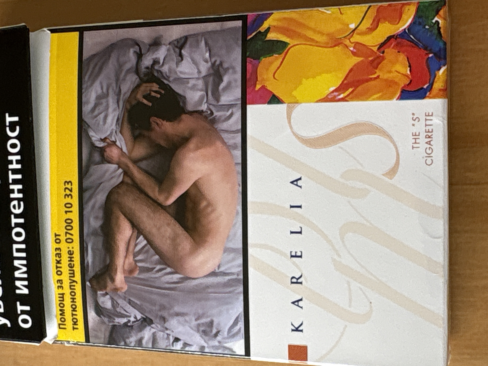 The image shows a close-up of a cigarette pack. The top of the pack has black text on a white background, with the words "от импотенция" visible. Below this is a yellow strip with black text that reads "Помощ за отказ от тютюнопушене: 0700 10 323". The main image on the pack is a photograph of a person lying in bed, covered by a grey sheet. The lower portion of the pack has a white background with a stylized "S" and the text "KARELIA" above it. To the right is a colorful abstract design. The bottom of the pack has the text "THE "S" CIGARETTE". The pack is placed on a wooden surface. The image does not contain any information about the city of Plovdiv, Bulgaria.