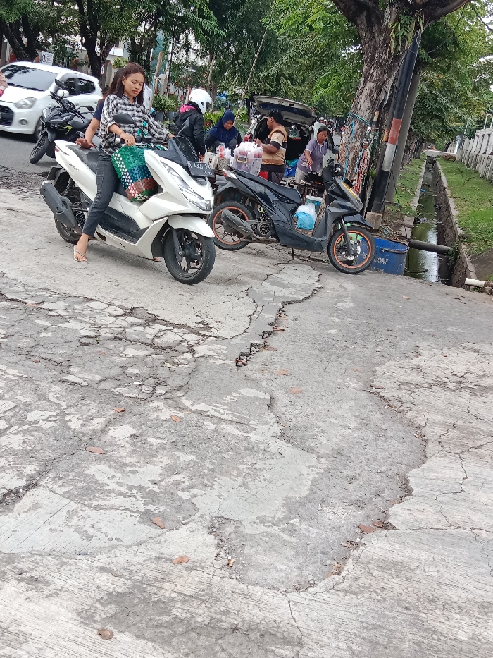A woman in a plaid shirt and blue jeans is riding a white scooter on a cracked, gray concrete road in Tugu, Indonesia. She is wearing a helmet and a green and white bag is hanging on the back of the scooter. There is a large tree on the right side of the road and a ditch with grass and water on the side of the road. There are four other people and two more scooters in the background, but they are out of focus. The woman is the primary focus of the image. The location of the scene is Tugu, Indonesia, which is located at the coordinates -6.973 latitude and 110.326 longitude. The time of day is daytime, and the weather is sunny.