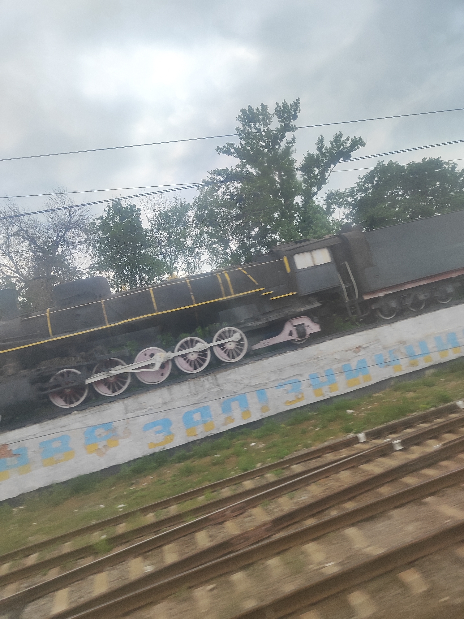 This is a full-shot, eye-level, medium-angle, blurry photograph of a black steam locomotive on display next to railroad tracks. The locomotive is dark gray with yellow and black trim. It has large spoked wheels with pink accents. In the background, there are trees and a cloudy sky. A concrete wall with blue and yellow painted letters is in front of the train. The image is taken from a moving vehicle, as indicated by the motion blur. The location is Unknown, Unknown.