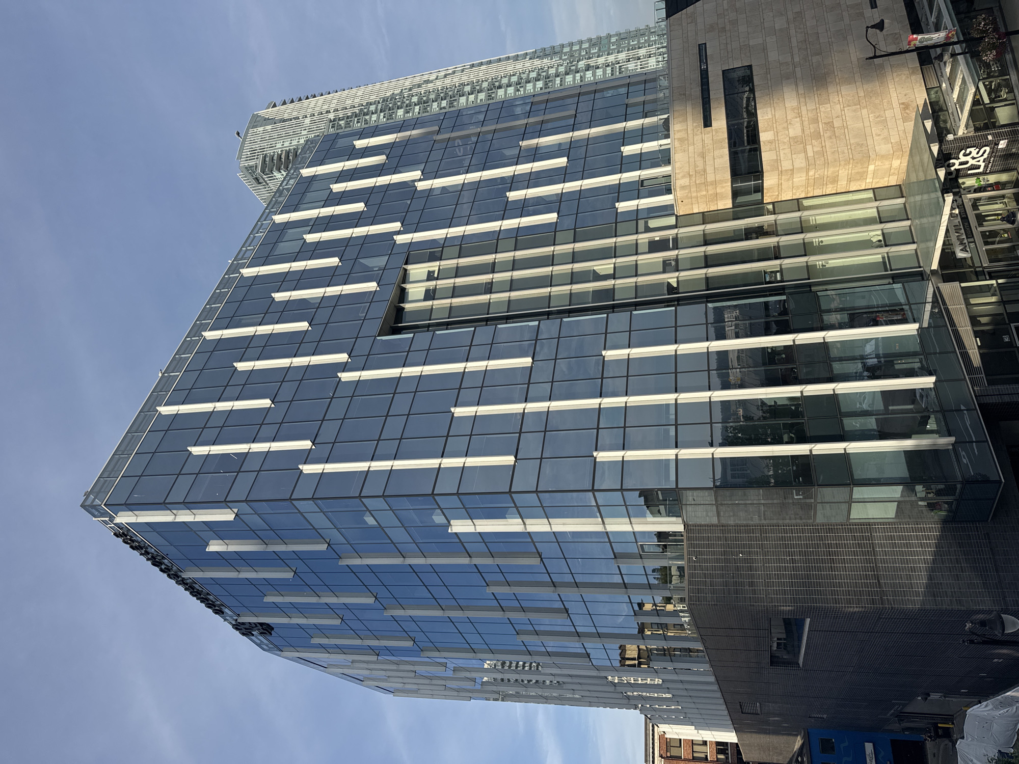 This is a photograph of the Douglas College building located in New Westminster, Canada. The primary subject is the modern, multi-story building with a glass facade. The building features vertical white accents against the blue glass panels. The bottom portion of the building is clad in dark brickwork, and a section to the right is faced with light-colored stone. The entrance area displays signage for "ANVIL" and "DOUGLAS COLLEGE". A taller, contemporary skyscraper is visible in the background to the right. The sky is a clear blue with some wispy clouds. The photograph appears to be taken from an elevated position, looking upwards at the building.
