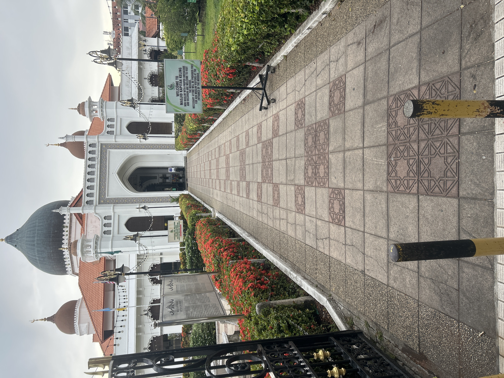 The image shows the entrance to the Kapitan Keling Mosque in George Town, Penang, Malaysia. A paved walkway, made of patterned square tiles, leads from the foreground towards the mosque's entrance. On either side of the walkway are neatly trimmed hedges with vibrant red flowers. To the left, a black ornate gate is visible, and behind it, informational signs are mounted on metal stands. To the right, a green sign on wheels, with the mosque's name and visitor information, is positioned near the walkway. The mosque itself is a white building with distinctive domes and minarets, featuring arched doorways and ornate architectural details. The sky is overcast. The scene is empty of people.