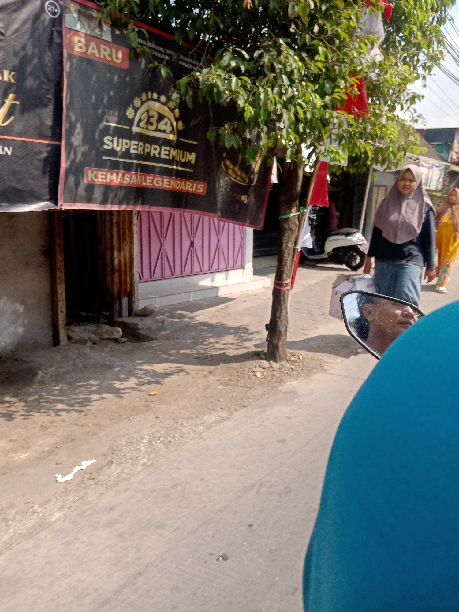 In Sugihwaras, Indonesia, on a sunlit day, a street scene unfolds. A black banner, likely advertising a product or event, hangs prominently. Text on the banner includes "BARU," "234 SUPERPREMIUM," and "KEMASANLEGENDARIS." A small tree with green leaves stands near the banner, adorned with red and white Indonesian flags. Two women are visible: one wearing a hijab and a dark top with blue jeans, the other in a yellow dress. A white scooter is parked behind them. In the foreground, a motorcycle's side mirror reflects a person's face. The ground is a mix of dirt and paved surface, with some scattered debris including a crumpled white object. The architecture suggests a commercial or residential area, with a building featuring a pink decorative gate visible in the background.