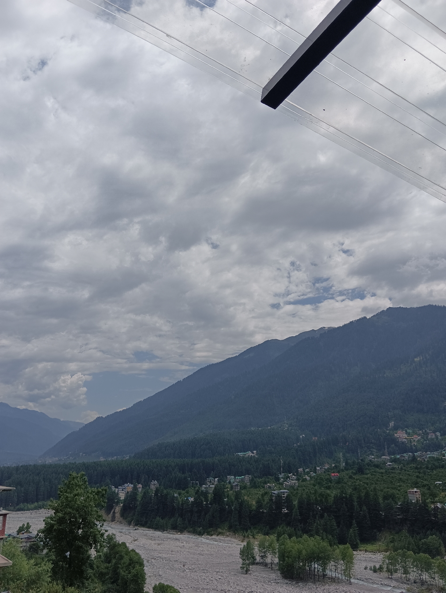 The image depicts a mountainous landscape under a cloudy sky. In the foreground, a rocky riverbed is visible with scattered trees. A dense forest covers the slopes of the mountains in the mid-ground, with a small settlement interspersed within the trees. The background features towering, tree-covered mountains. A portion of a transparent structure with dark horizontal elements is visible in the top part of the image, suggesting an elevated vantage point. The overall scene is natural and serene, possibly capturing a view of Vashisht, India, known for its scenic beauty and mountainous terrain.