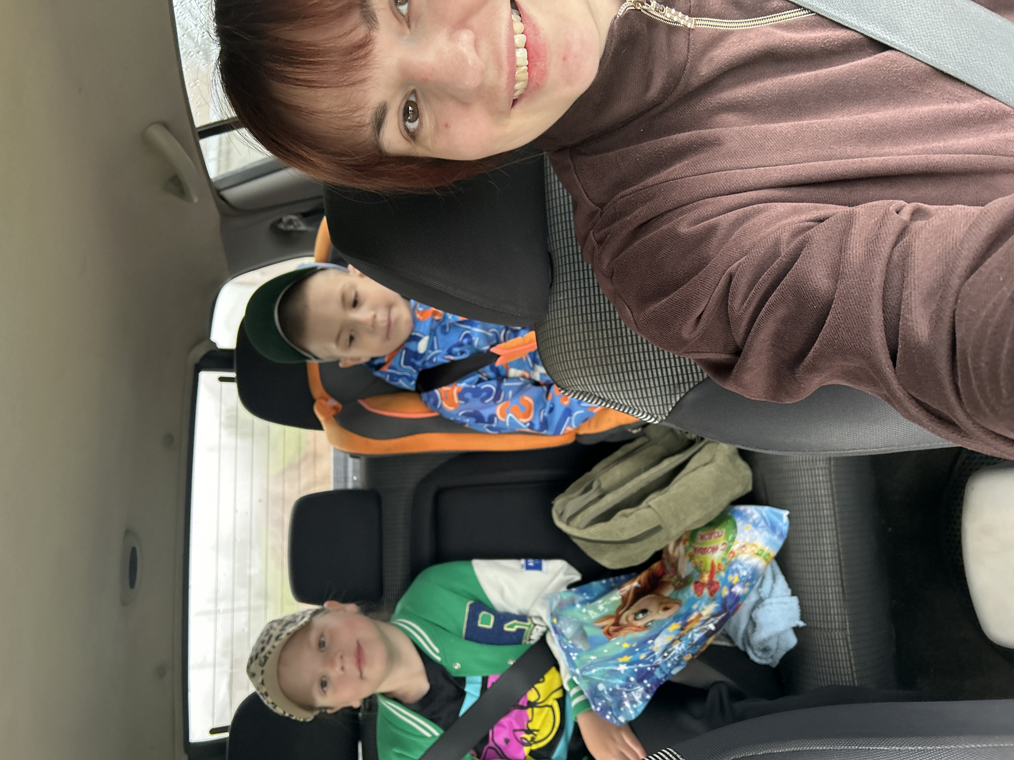 The image is a selfie taken inside a car, featuring a woman and two children in the back seats. The woman, with reddish-brown hair and a wide smile, is in the foreground, wearing a brown velour-like zippered top and a seatbelt across her chest.
Behind her, a young boy is secured in a child car seat. He wears a blue and orange patterned long-sleeved shirt and a green baseball cap, looking forward or slightly down. In the rearmost visible seat, a young girl is seated, wearing a green and white varsity-style jacket, a colorful t-shirt, and a leopard-print cap. She looks towards the camera with a gentle smile, also wearing a seatbelt.
The car's interior features grey and black seats and headrests, with a light-colored ceiling. Through the car windows, an overcast sky is visible, with some streaks on the glass suggesting rain or condensation, indicating a cloudy daytime. On the seat next to the girl, a green bag is visible, along with a blue blanket or towel featuring the Disney character Elsa from "Frozen." Partial Russian text on this item clearly spells out "С НОВЫМ ГОДОМ!" (Happy New Year!). A white container is also partially visible on the floor or seat in the lower right. The scene depicts a casual journey or outing in a vehicle, likely within Фоки, Russia, given the context. The overall mood is calm and familial.