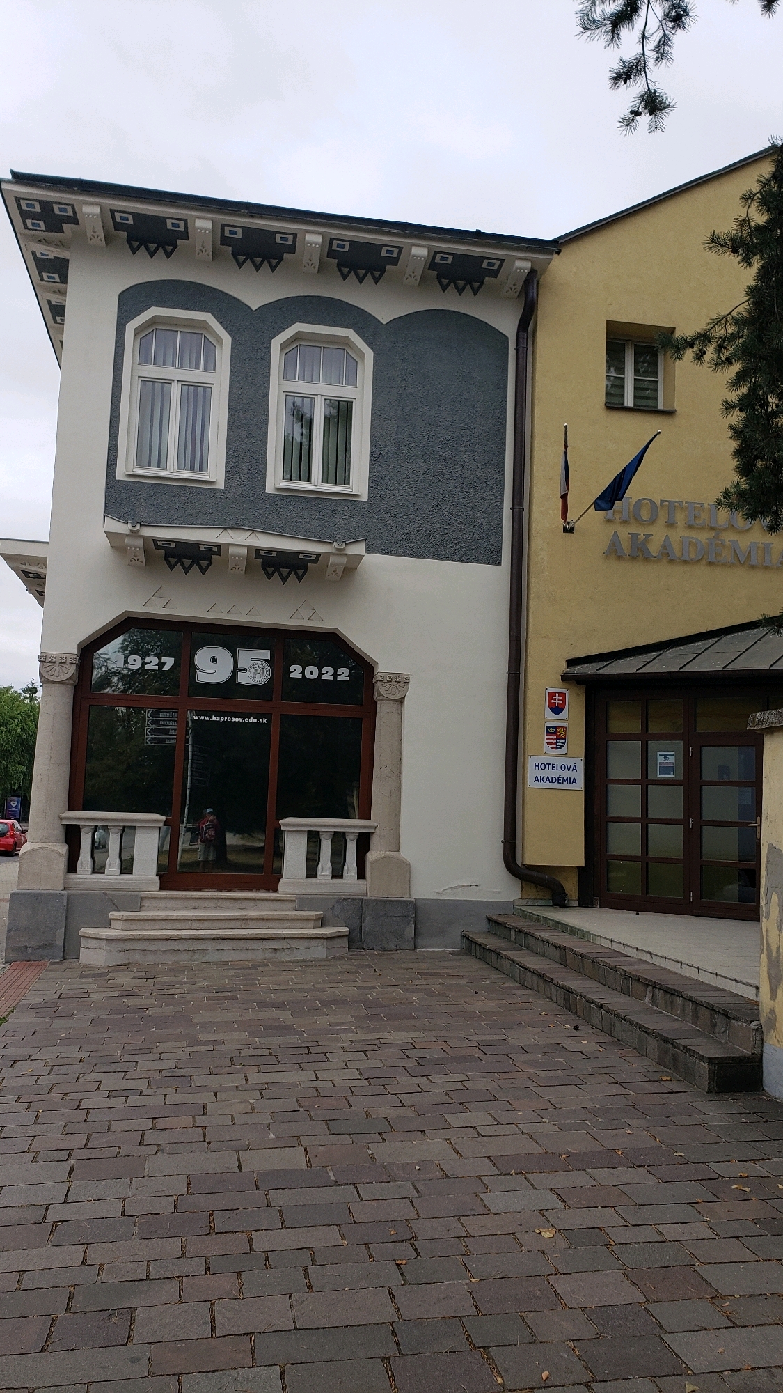 The image shows the exterior of the Hotelová Akadémia in Prešov, Slovakia. The building features a striking contrast between two architectural styles. On the left, a light-colored facade with dark grey decorative elements and arched windows is connected to a pale yellow building on the right. The yellow building has a simpler design and houses the hotel entrance.
A large window on the left section of the building displays "1927 95 2022" and the website "www.hapresov.edu.sk," indicating an anniversary or significant milestone. Below this window, a staircase leads to a dark-framed glass door. The yellow building on the right has a smaller window, a flag with the European Union and Slovak Republic emblems, and a sign that reads "HOTELOVÁ AKADÉMIA." Two sets of stone stairs lead up to the entrance of this section. The foreground is paved with dark grey rectangular stones. The sky is overcast, suggesting a cloudy day. There are no people clearly visible in the scene.