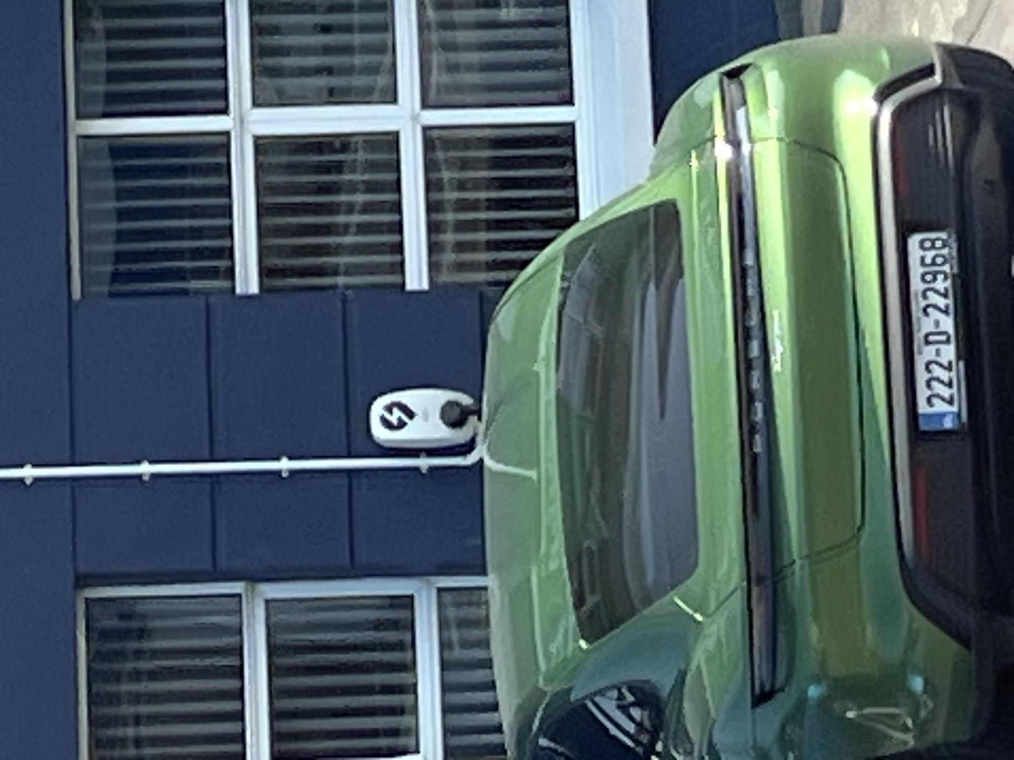 A green electric car, possibly a Porsche Taycan, is parked in front of a dark blue building with barred windows. A white electric car charging station is mounted on the building wall to the right of the windows. The car's license plate is visible and reads "22-D-22968". The image appears to be taken in an urban setting.