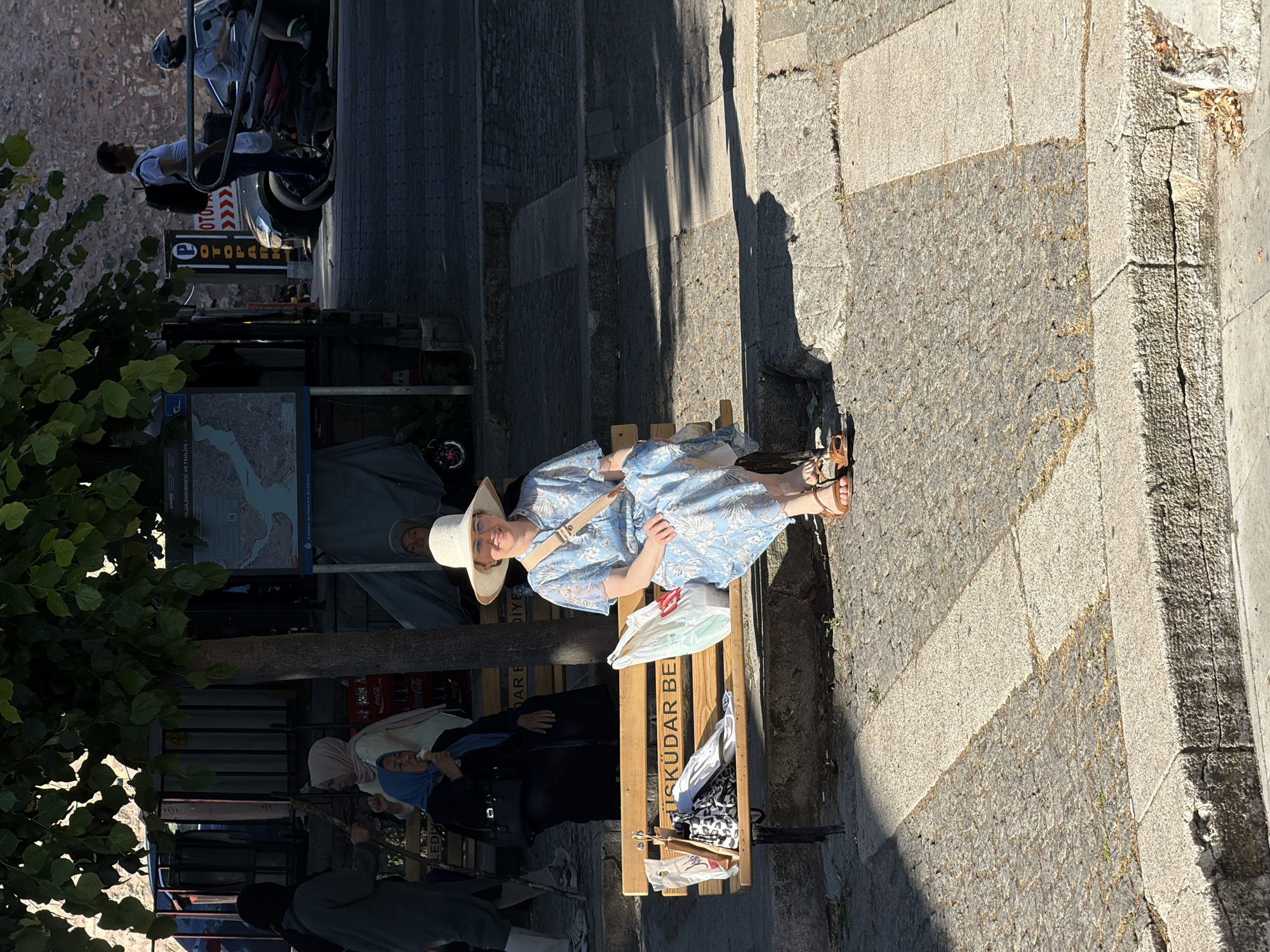 In Üsküdar, Turkey, a woman wearing a white sunhat and a light blue floral dress sits on a wooden bench. The bench has "ÜSKÜDAR BE" inscribed on it. Next to her are several shopping bags and an umbrella. To her left, another woman in a blue headscarf eats ice cream. In the background, other people are visible, along with a map displaying a waterway. The scene is set on a sunlit street with cobblestone paving.