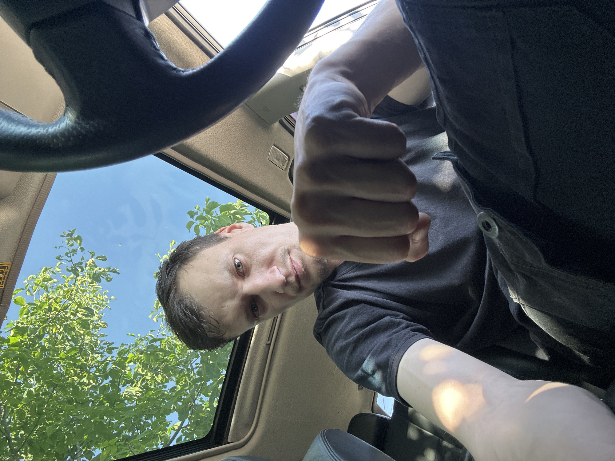 A man is inside a car, his fist is extended towards the camera, and he is looking at the camera. The sunroof of the car is open, and green leaves and a blue sky are visible through it. The man is wearing a black t-shirt and black pants. He appears to be of Caucasian descent, with short brown hair and a light beard. The interior of the car is visible, including the steering wheel and part of the dashboard. The lighting suggests it is daytime. The image was taken in Shatava, Ukraine.