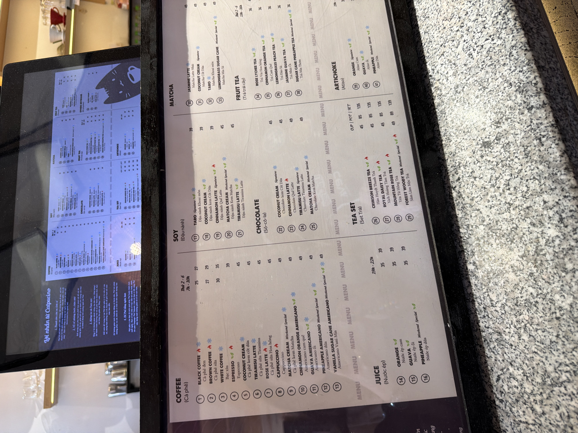 The image shows a menu board, likely from a café in Quy Nhon Ward, Vietnam. The menu features a variety of drinks including coffee, soy milk based beverages, matcha, fruit teas, chocolate drinks, juices, and artichoke tea. The price list is displayed next to each menu item. Some items are labeled as "Weekend Special" or "Signature." The upper left corner of the image shows a tablet displaying the same menu.