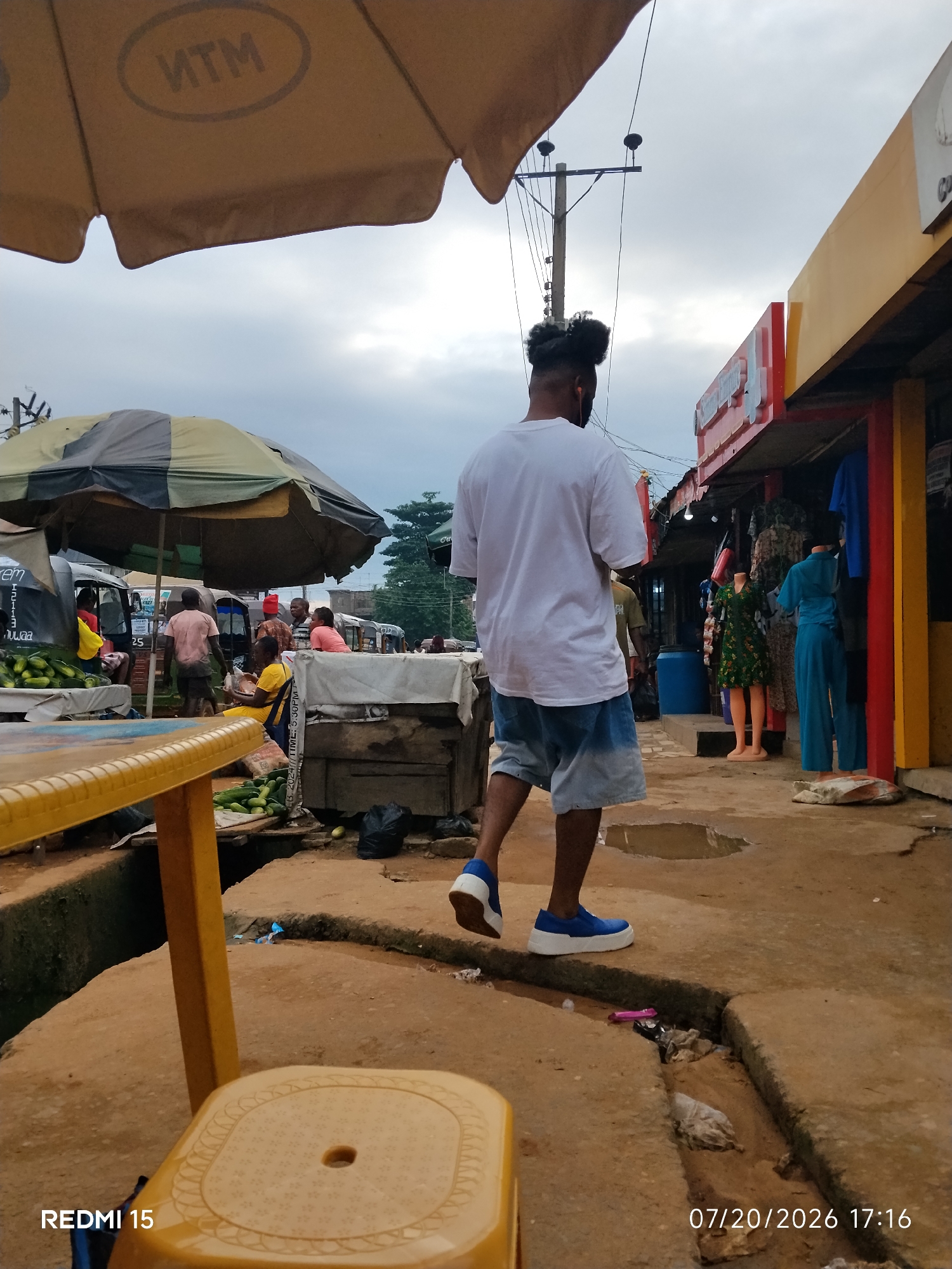 The image depicts a street scene in Umuode, Nigeria. In the foreground, a yellow plastic chair and table are partially visible. A man in a white t-shirt and denim shorts is walking away from the camera, his back to the viewer. He is wearing blue sneakers with white soles. To his left, there are market stalls with various goods, including cucumbers and clothing displayed on mannequins. A yellow and red awning extends from a building on the right, and a power pole with electrical wires is visible in the background. The ground appears to be unpaved and has a small drainage ditch filled with debris. The sky is overcast.