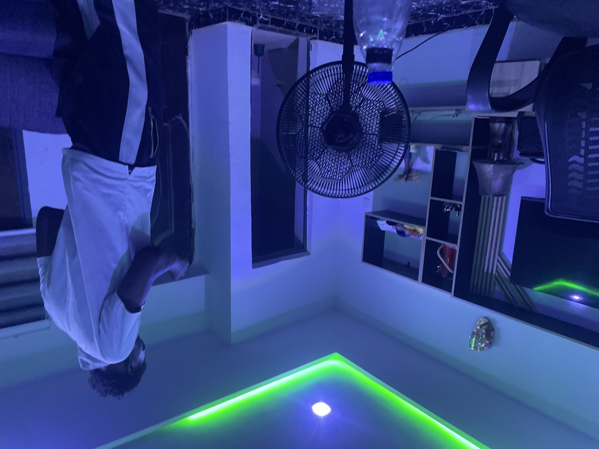 The image portrays an indoor room, likely a contemporary living or bedroom space, dominated by artificial lighting. The walls are light-colored, possibly white, and are bathed in a strong blue ambient light, suggesting an evening or nighttime setting.
On the left side of the frame, a person is visible from the waist up. They have dark, curly hair and are wearing a white t-shirt. Their lower half is clad in dark trousers featuring a distinct white stripe along the side. The person's head is tilted slightly upwards, facing generally towards the viewer.
Key objects within the room include a black wall-mounted oscillating fan, prominently featured in the upper central area. Above the fan, a clear plastic water bottle with a blue cap is visible, possibly suspended or part of a makeshift setup. To the right, a dark shelving unit is visible, holding various indistinct items, and a portion of a black office-style chair is also present. In the background, there's an opening or window with a dark frame.
The ceiling features a modern lighting design, with a prominent L-shaped perimeter of vibrant green LED strip lighting. A single bright white spotlight is embedded centrally within this green-lit area. A small, reflective object, possibly decorative, is mounted on the wall near the green LED light.
The scene conveys a cool, modern aesthetic due to the intense blue and green artificial illumination. No specific activity beyond the person's presence is discernible. There is no visible text in the image. The location context for this scene is Abuja, Nigeria.