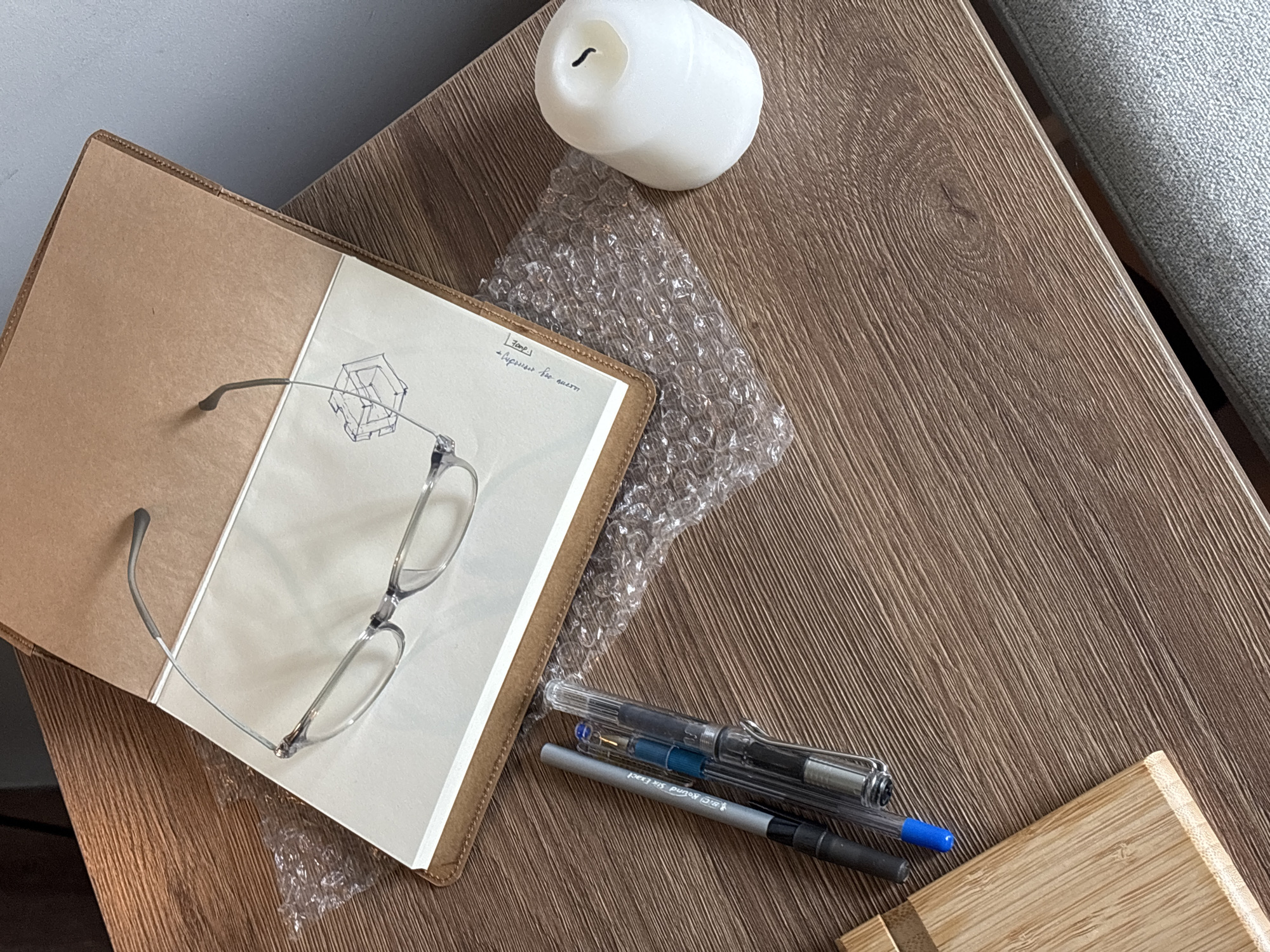 The image shows a still life arrangement on a wooden surface. A notebook, opened to a blank page with a geometric drawing and some handwriting, sits to the left. A pair of eyeglasses rests on the open pages. A lit white candle sits at the top of the image. Bubble wrap is visible under the notebook and beside the candle. Two pens are positioned to the right of the notebook. A small wooden object is in the bottom right corner. The setting appears to be indoors. The location is not discernible from the image.