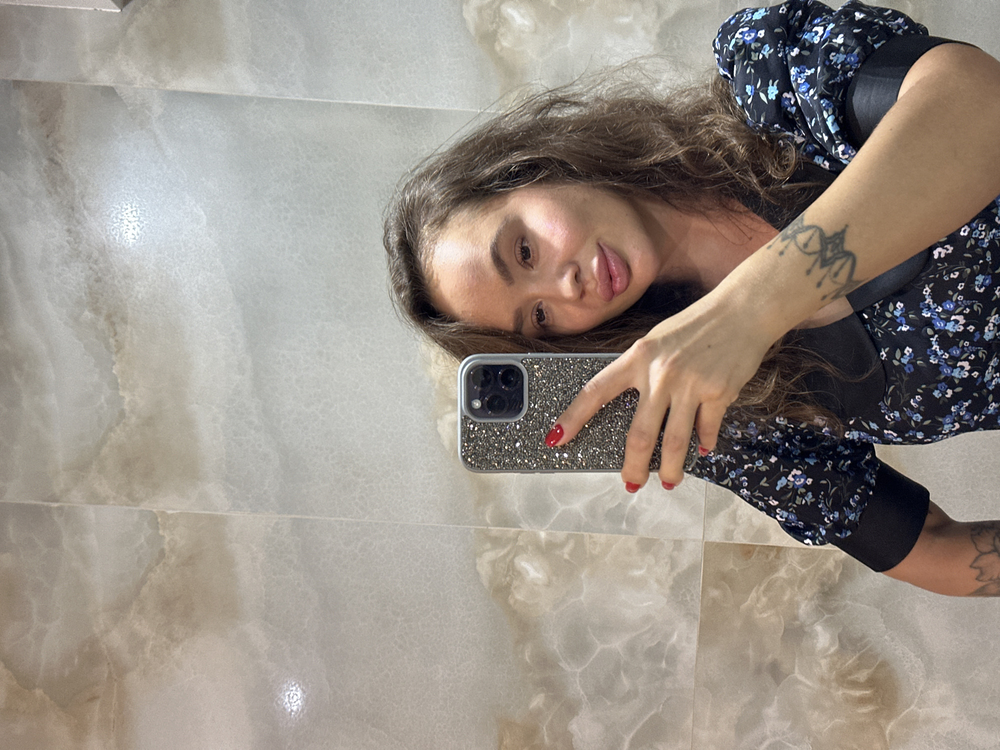 A woman is taking a selfie in a bathroom with marble-like wall tiles. She is wearing a black dress with a floral pattern and has long, wavy brown hair. She is holding a phone in her right hand. The scene is indoors, and the lighting appears to be artificial. There are no specific details that indicate the location in Duvankul', Russia.