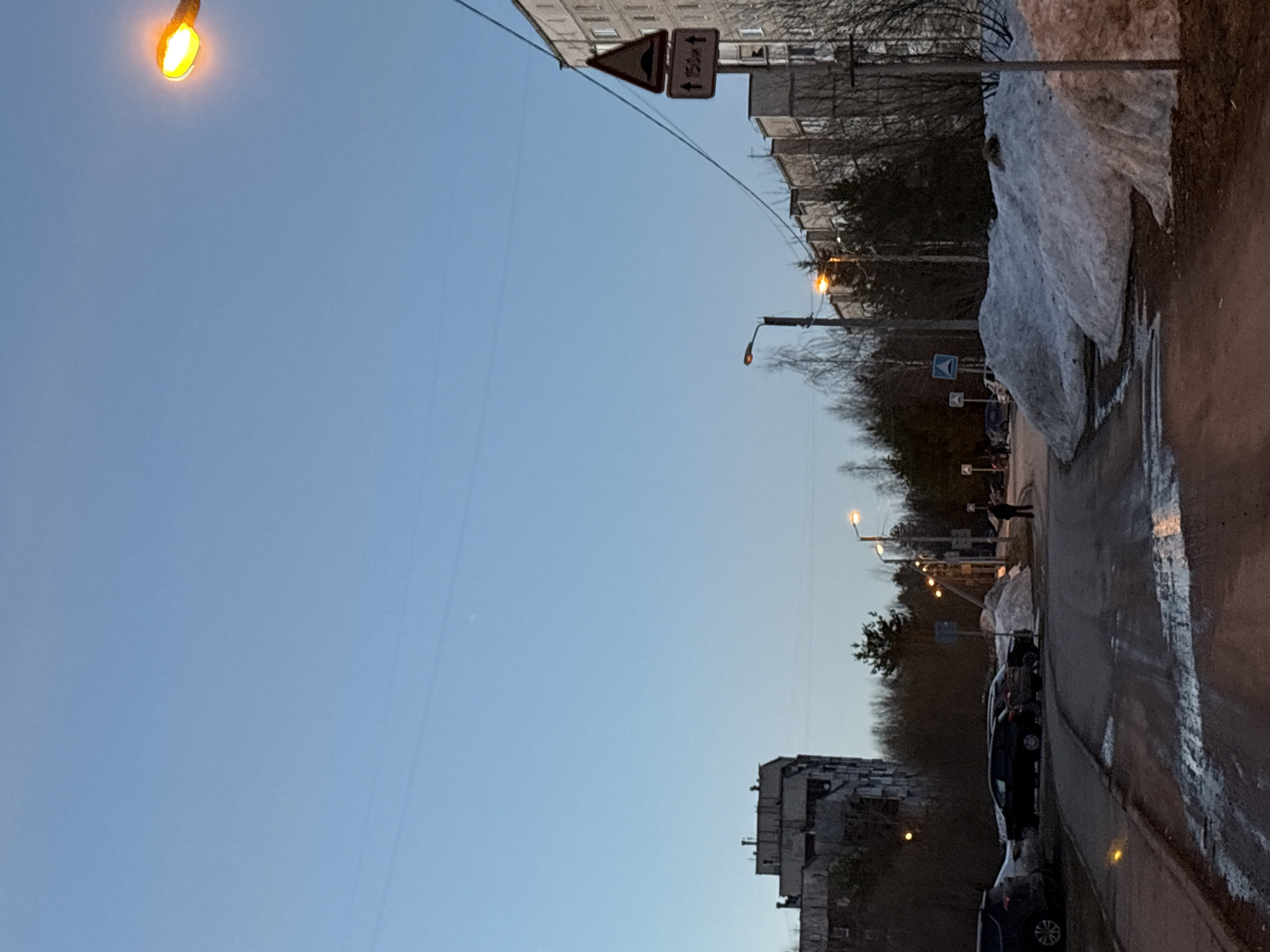 The image shows a street scene in Berezniki, Russia. The sky is a clear light blue, with a visible streetlight in the upper left corner. Buildings are visible along the sides of the street, with some appearing to be older apartment blocks. Along the right side of the frame, there is a sidewalk covered with melting snow and puddles of water. Several parked cars line the street. In the distance, there are people walking along the sidewalk. Street signs are mounted on poles near the buildings. Trees without leaves can be seen in the background.