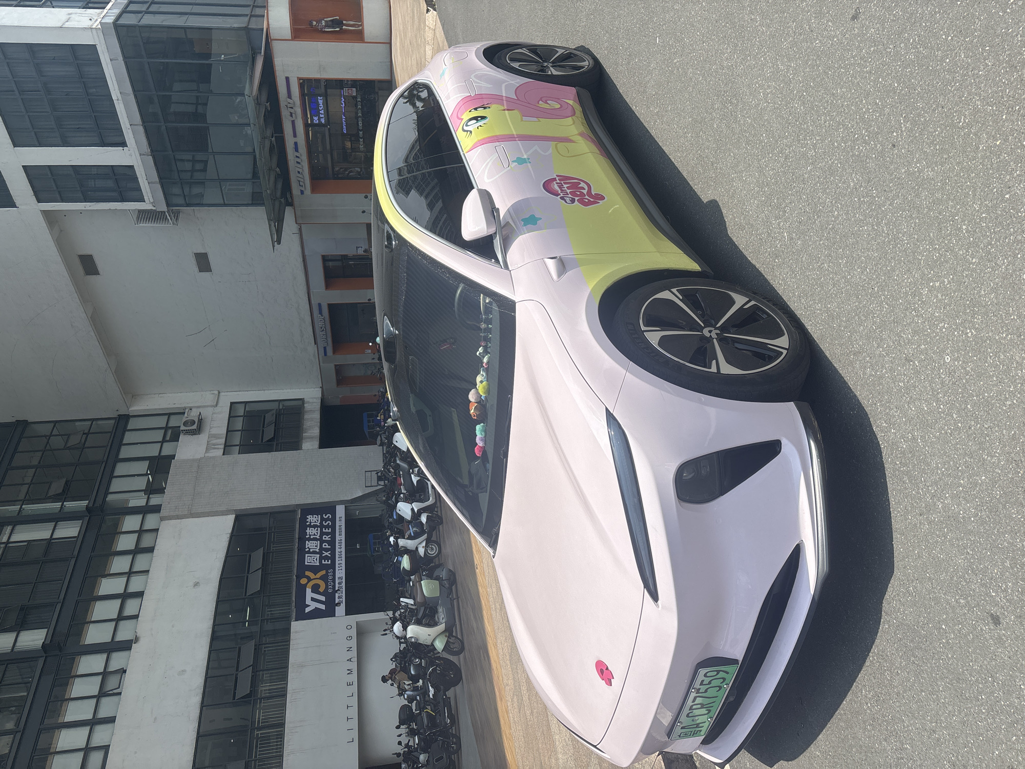 In Baiyun District, China, a pink and yellow electric car is parked on an asphalt road in front of a multi-story building. The car features a "My Little Pony" graphic on its side and a pink floral emblem on the hood. The building has a modern facade with large windows and some signage. A row of parked scooters is visible behind the car, and a few individuals are present on the sidewalk. The scene appears to be a bright, sunny day.