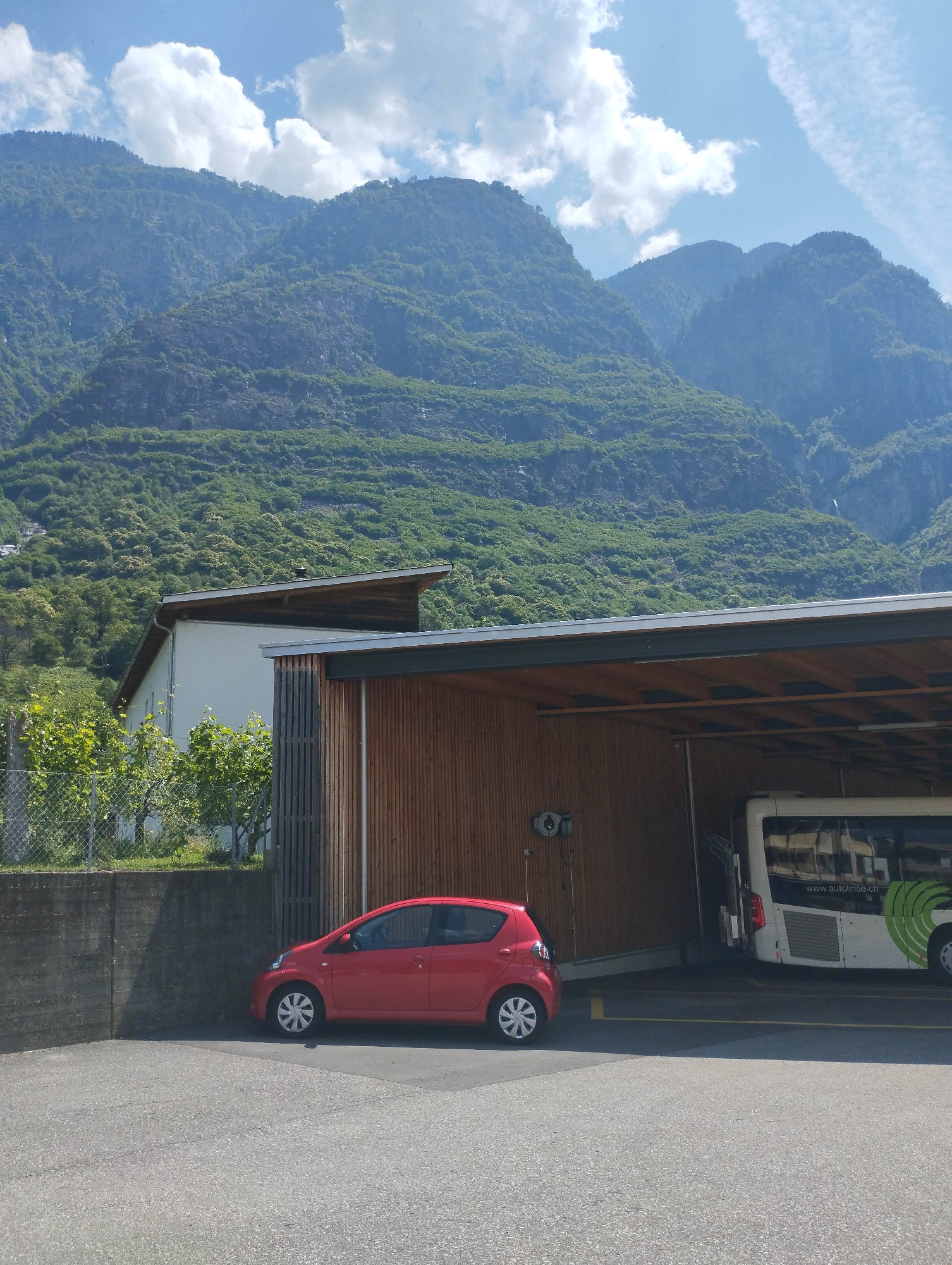 A red car is parked next to a concrete wall. Behind the car is a covered structure with wooden siding and a large bus parked inside. The structure is next to a white building. In the background, lush green mountains with rocky outcrops rise towards a blue sky with white clouds. The scene appears to be in Biasca, Switzerland, given the presence of a bus with "www.autolinee.ch" on its side, indicating a Swiss bus company.