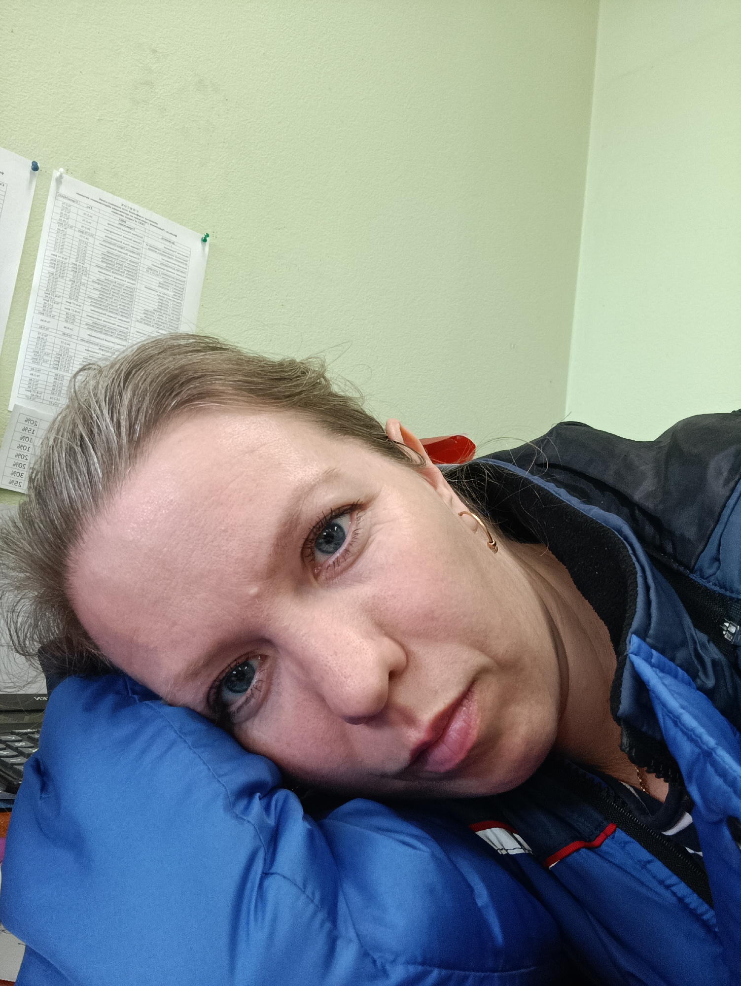 An adult female is captured in a close-up, angled shot, resting her head on her left arm or shoulder. She gazes directly at the camera with a neutral, perhaps contemplative or weary expression. Her hair is a mix of light brown and grey. She is dressed in a blue and black jacket, possibly a puffer style, over a striped undergarment. A small golden hoop earring adorns her right ear.
The setting appears to be indoors, likely in Chaikovsky, Russia, given the context. The background is dominated by a plain, light green wall. In the upper left portion of the frame, several sheets of paper are pinned to the wall with blue and white thumbtacks. These documents feature visible Cyrillic text, appearing to be lists or schedules, with the word "СПИСОК" (SPISOK), meaning "list," partially legible on one sheet. The arrangement suggests an office, classroom, or institutional environment. The lighting is consistent with an indoor setting, and no specific time of day or weather can be discerned.