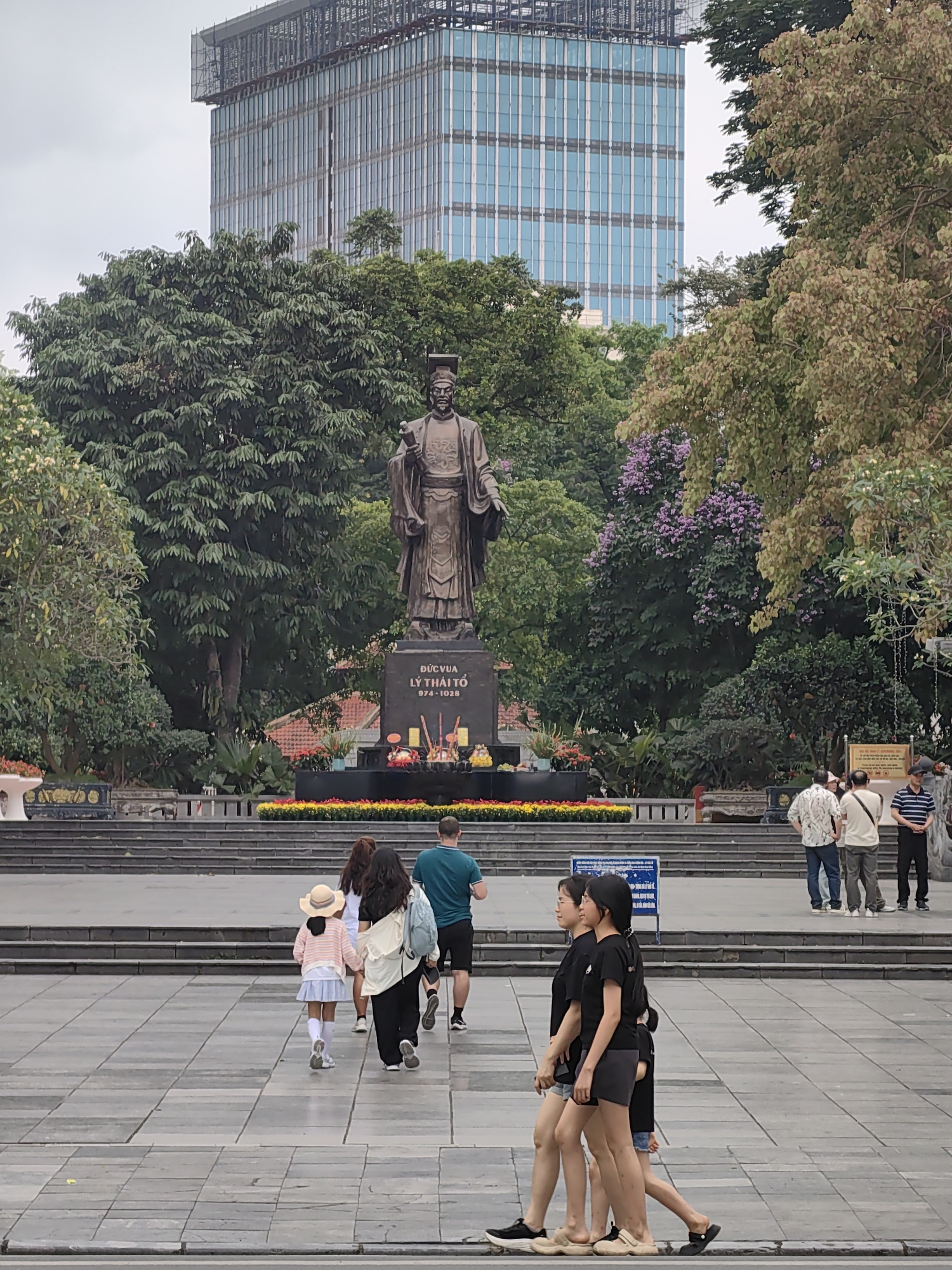This image depicts a public square or park in Hà Nội, Vietnam, centered around a large bronze statue of Emperor Lý Thái Tổ. The statue, showcasing the historical figure in traditional attire and holding a scroll, rests on a dark stone pedestal inscribed with "ĐỨC VUA LÝ THÁI TỔ 974 - 1028." A platform in front of the statue holds offerings of incense, candles, and flowers, bordered by bright yellow and red floral arrangements.
The monument is set amidst lush green trees and some flowering shrubs, with hints of purple blossoms. In the background, a modern high-rise building with a blue glass facade and scaffolding at its upper levels is visible, contrasting with the historical statue. The sky is overcast, indicating a cloudy daytime.
Several groups of people are present in the square. In the foreground, two young women in black t-shirts and shorts walk across the paved grey tiles. In the mid-ground, a family, including a young girl in a straw hat, walks towards the statue, while other individuals stand near the steps observing the scene. The overall impression is one of a public space where history and modernity intertwine, attracting visitors.