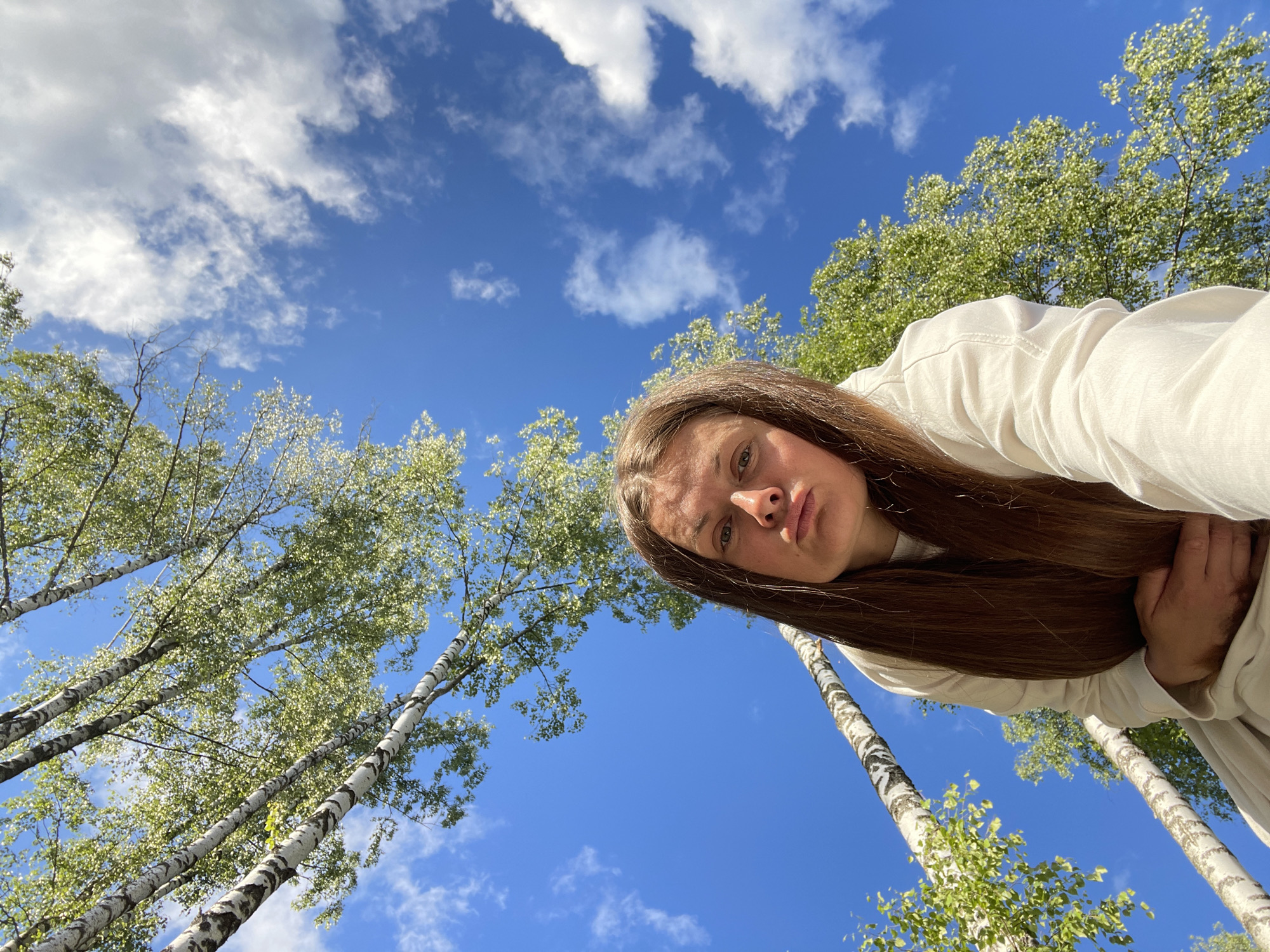 The image shows a low-angle, wide shot of a person outdoors on a bright, sunny day. The person, who appears to be a young woman with long brown hair, is looking up towards the camera, their face partially visible at the top right of the frame. They are wearing a light-colored garment, possibly a hoodie or sweatshirt.
The setting is a forest with tall birch trees, their white bark with black markings characteristic of the species. The trees are full of vibrant green leaves, suggesting a time of year like late spring or summer. The sky is a clear, deep blue, dotted with scattered white, fluffy clouds. The perspective, looking up from a low angle, emphasizes the height of the trees and the vastness of the sky.
The overall mood is serene and natural, with the bright sunlight and lush greenery creating a pleasant atmosphere. There is no visible text in the image. The location context provided is Zarechny, Russia. The activity appears to be a casual moment of enjoying nature, possibly taking a selfie or simply looking up at the sky.