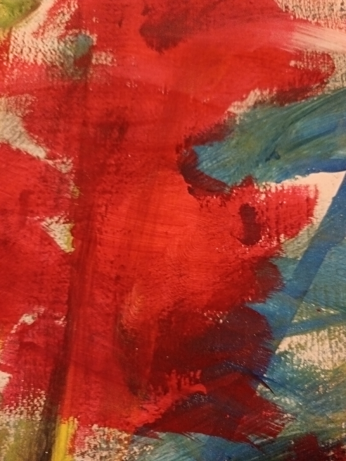 The image displays a close-up view of an abstract painting characterized by broad, textured brushstrokes. The dominant color is red, applied in thick, sweeping motions across the central and left portions of the composition. Interspersed with the red, particularly on the right and bottom right, are areas of teal and blue paint, also exhibiting visible brushwork. Patches of a lighter, possibly white or off-white, background material are visible where the paint is less dense, revealing the underlying surface. Minor accents of yellow and green appear faintly on the far left. The painting's texture suggests the use of acrylic or oil paints. There are no identifiable people, specific objects, or discernible actions within the artwork. The image itself provides no visual information to determine its specific location within the city of Kamenz - Kamjenc, Germany, or any other geographical point.