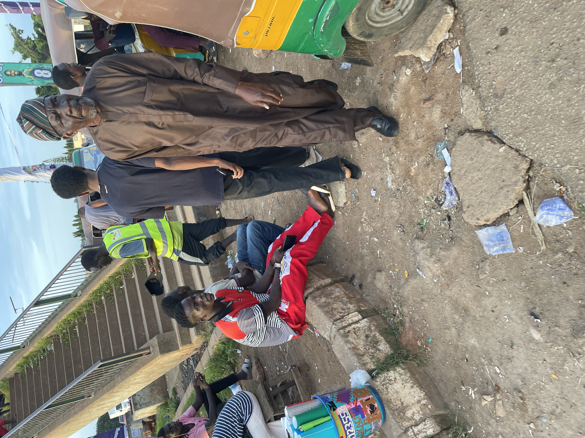 A group of men are gathered on a dusty, debris-strewn roadside in Gura Top, Nigeria, near an overpass. In the foreground, a young man in red and black striped shirt and red pants sits on a concrete curb, holding a phone. To his right, an older man in a brown traditional tunic and cap stands, looking forward. Behind him, other men are seen standing and sitting. A colorful tricycle, labeled "PETROL," is parked nearby. The overpass, made of concrete stairs, is visible in the background, with some vegetation growing along its sides. The sky above is a clear blue with some clouds.