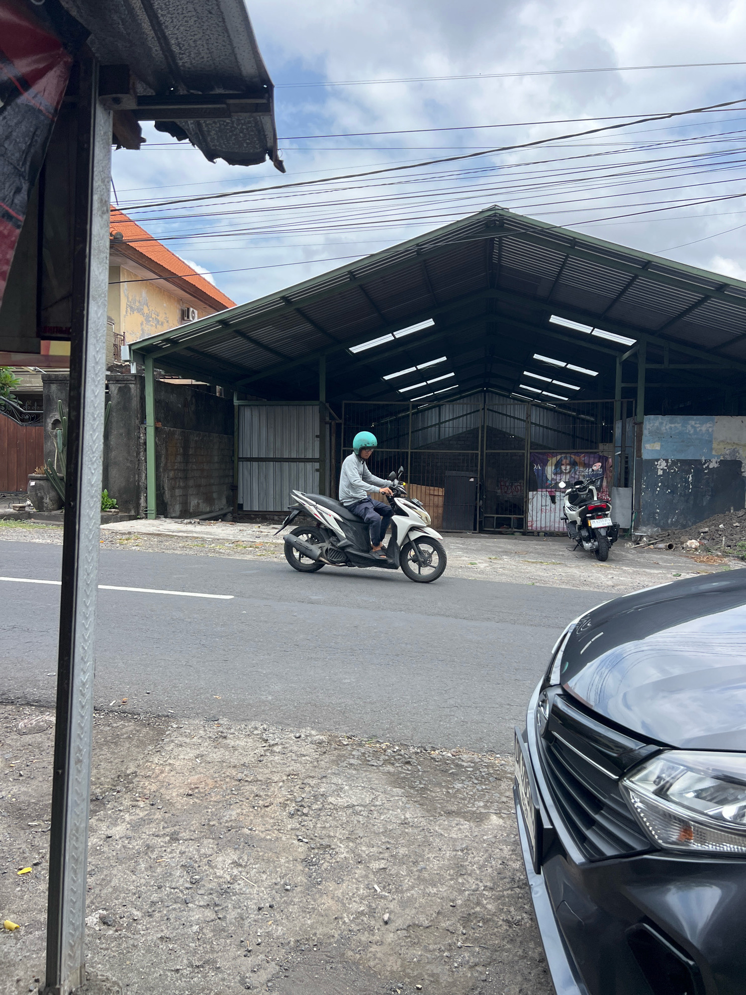 A person wearing a blue helmet and a grey long-sleeved shirt rides a white scooter on a paved road in Denpasar, Indonesia. The scooter is moving from left to right. In the background, there is a large, open-sided building with a corrugated metal roof. To the left of the building, there is a residential building with an orange tiled roof and a yellow, weathered wall. A dark-colored car is parked in the foreground on the right side of the image. The sky is cloudy.
