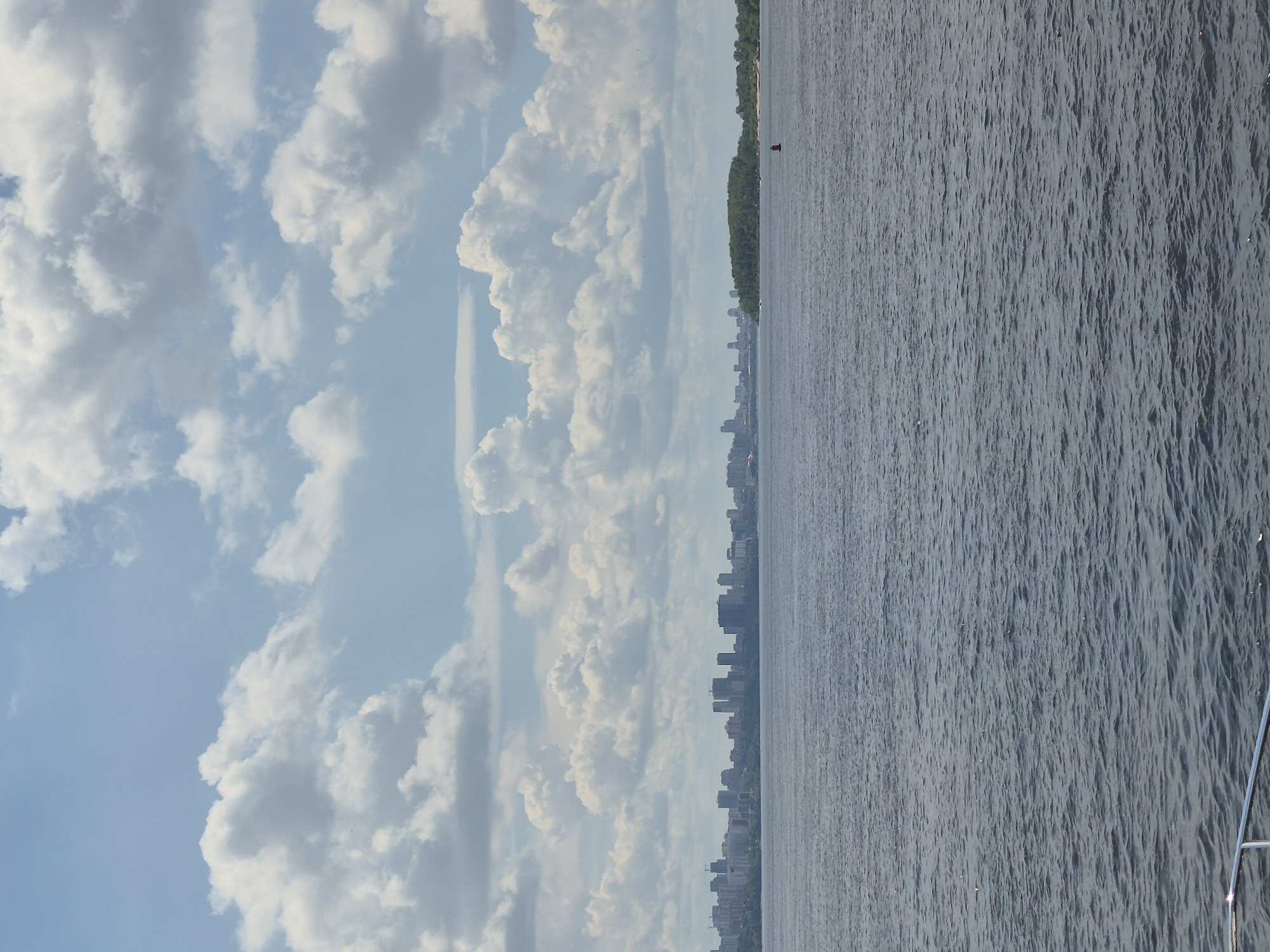 The image shows a wide river with a distant city skyline on the horizon. The sky is filled with fluffy white clouds against a light blue background. The river water is dark and appears choppy with small waves. The city skyline is composed of numerous buildings, including some tall structures that suggest a modern urban area. On the right side of the frame, there is a green, tree-covered island with a small sandy area and a red buoy in the water. The foreground shows a metal railing, suggesting the photo was taken from a boat or a pier. The overall impression is a sunny day over a large body of water with a city in the background. This scene is consistent with the view of the city of Samara, Russia, across the Volga River, with the city's embankment and skyline visible in the distance.