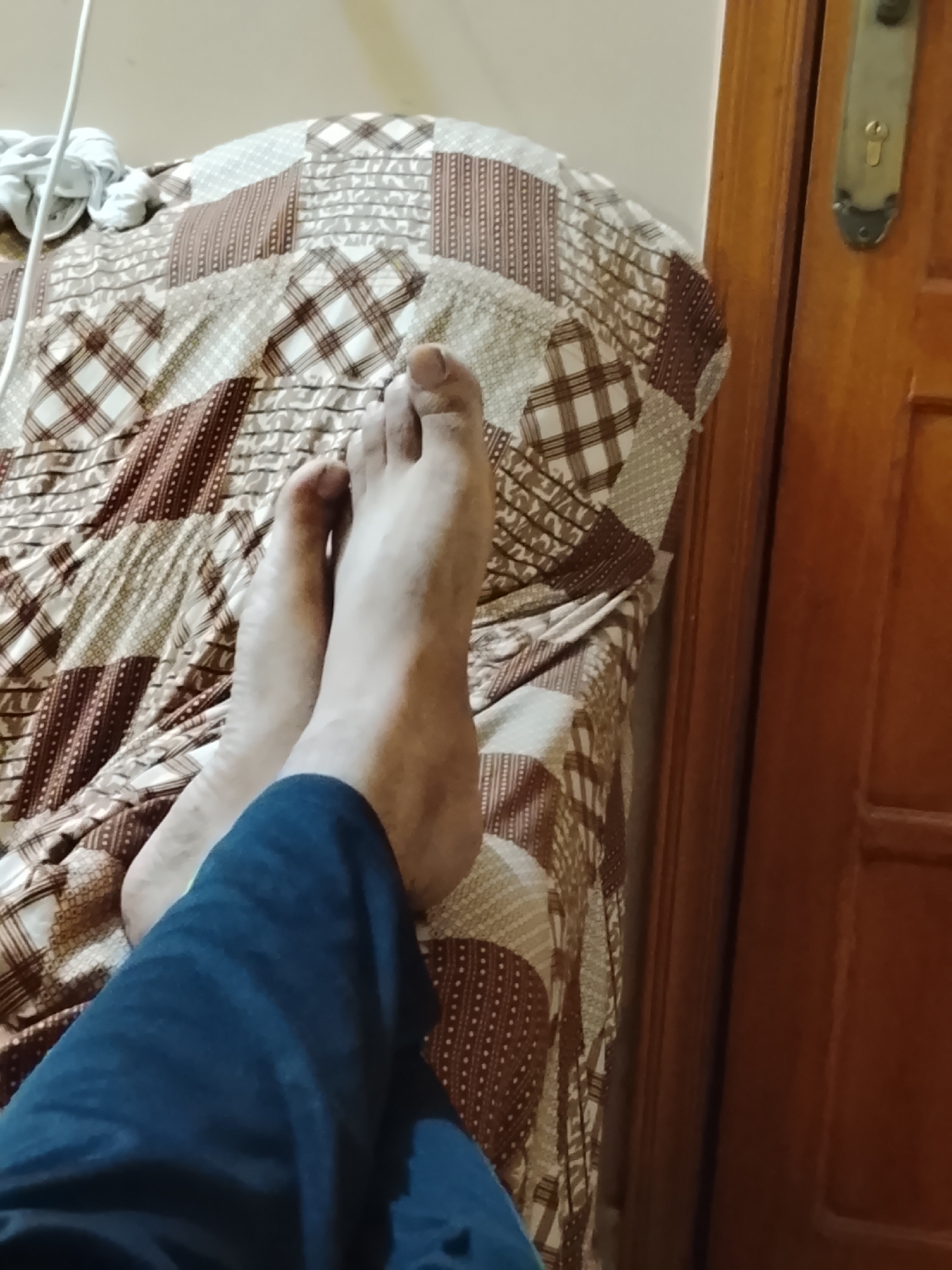 The image shows a pair of bare feet resting on a bed covered with a patterned quilt. A portion of a person's blue pants is visible in the foreground. To the right, a brown wooden door with a metal handle is partially visible. The location is assumed to be a domestic interior, city of Multan, Pakistan.