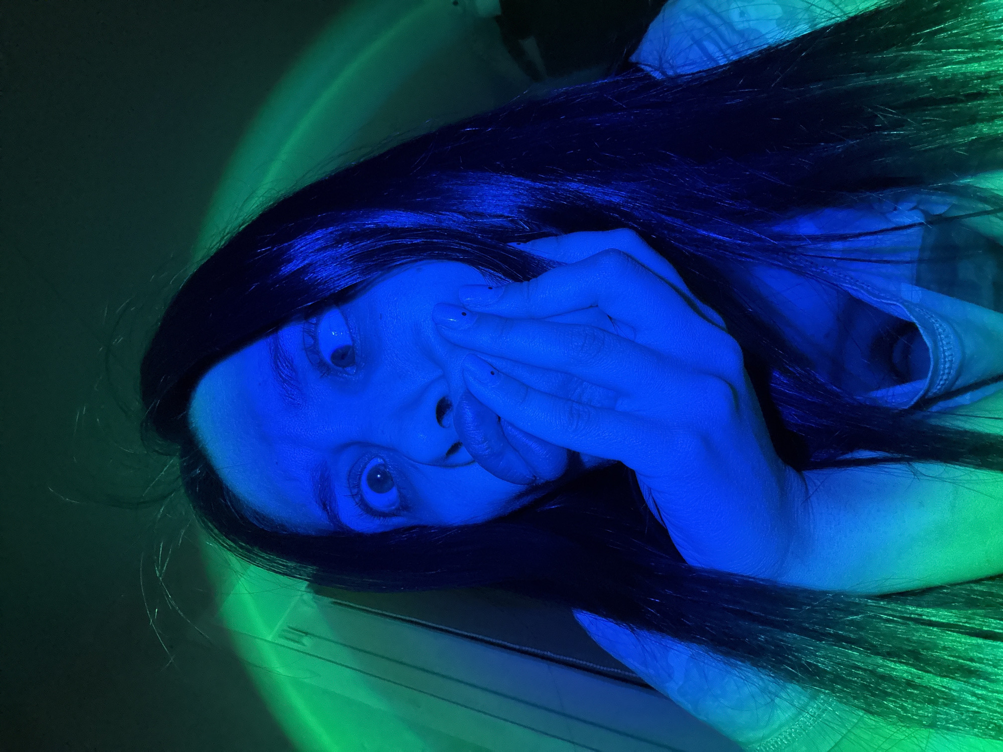 A woman with long, dark hair is lying down, angled with her head toward the lower left corner. Her face and hands are illuminated with blue light, while the surrounding areas are lit with green. Her right hand is partially covering her mouth. The location is not discernible.