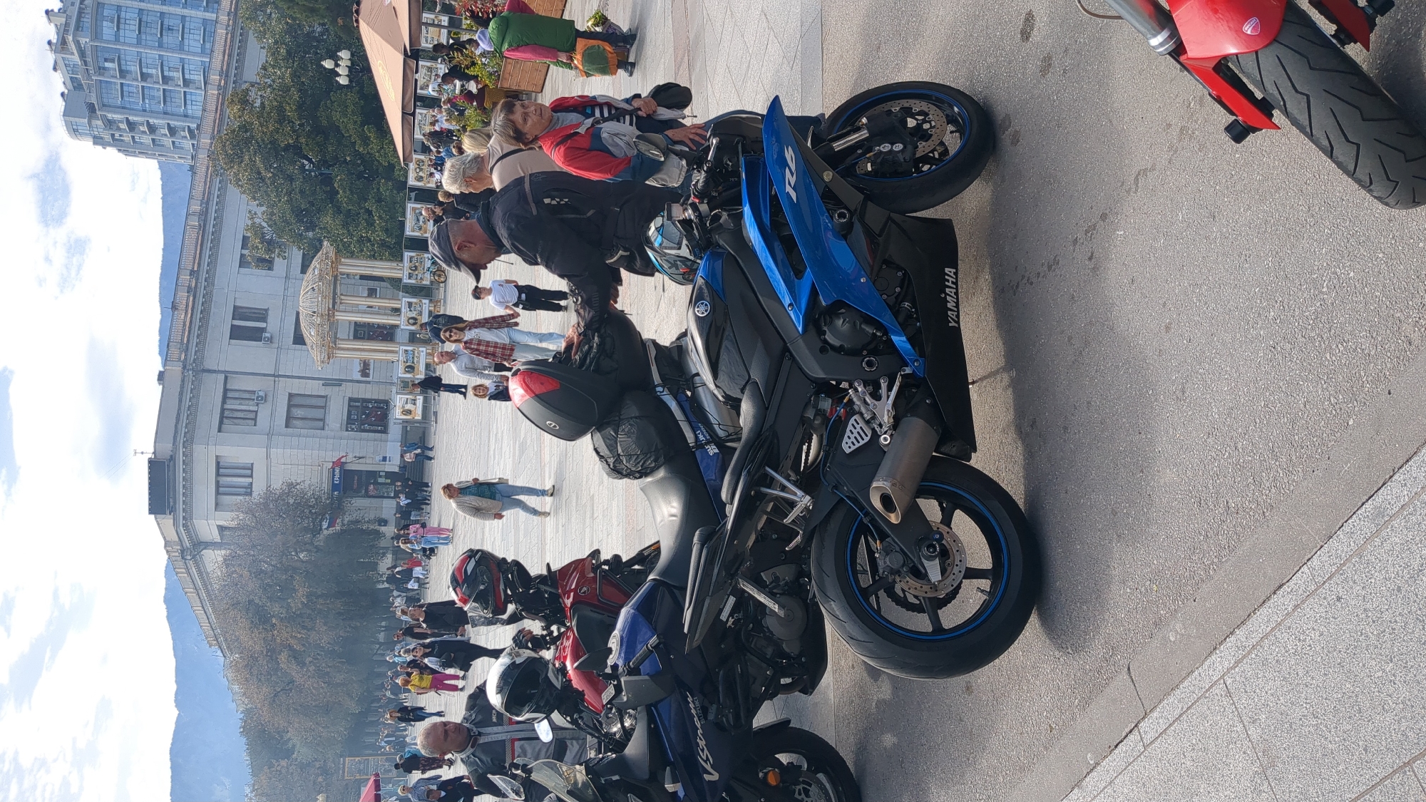 The image shows a street scene in the city of город Ялта, Ukraine. Several motorcycles are parked along the edge of the street, with people walking by and sitting at tables. A blue Yamaha motorcycle is prominently featured in the foreground. In the background, there are buildings and trees. There is a building that appears to be the local City Hall. There are mountains in the background.