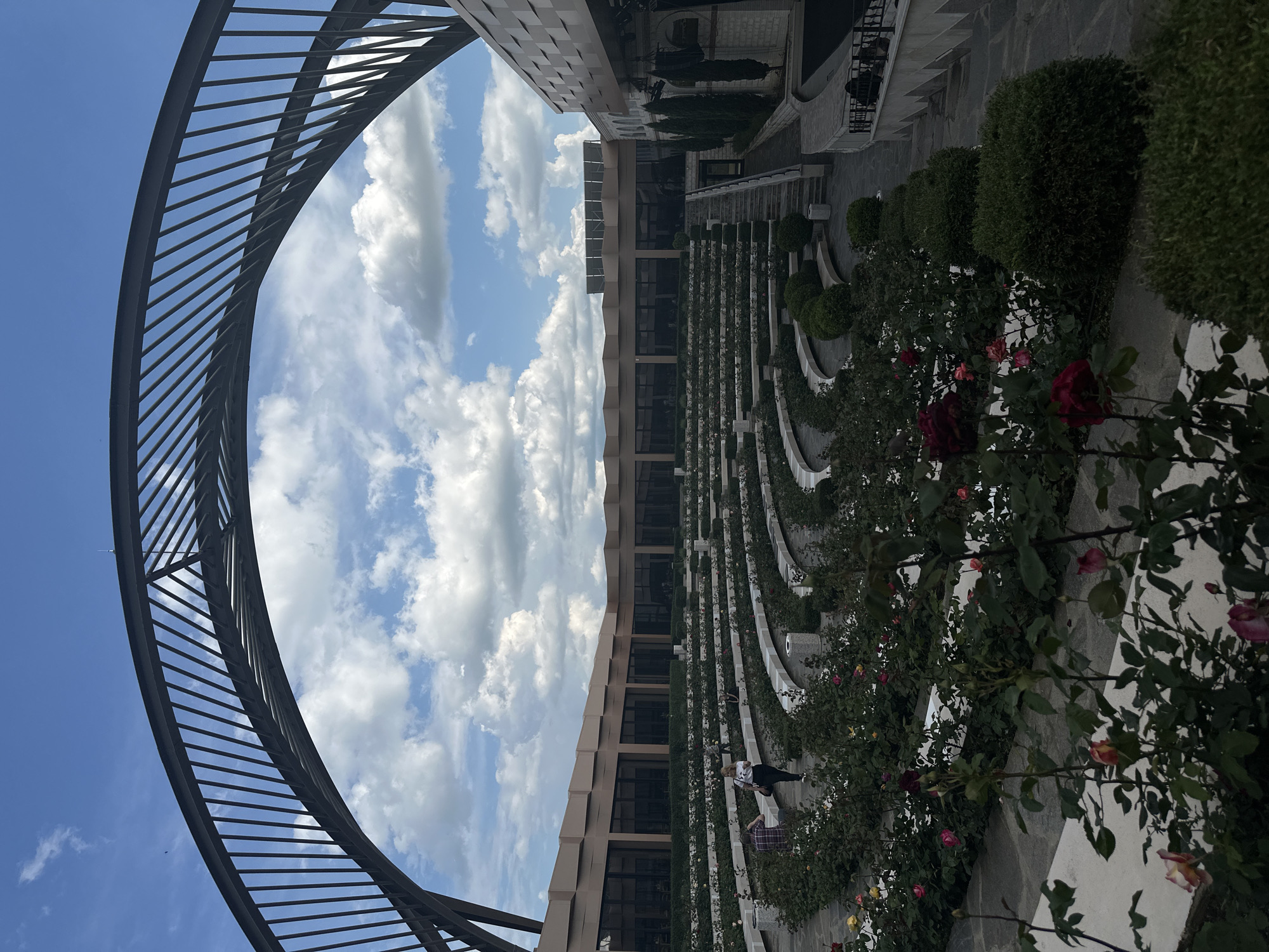 The image, taken in Skobelevo, Bulgaria, shows an outdoor scene featuring a circular metal structure in the foreground, framing a view of the sky with scattered clouds. To the right, a tiered garden extends into the distance with rows of roses and small bushes. At least two people are on the garden path. A building with a modern design is also visible, partially reflecting the sky. The scene is captured from a low angle, emphasizing the height of the metal structure and the tiers of the garden.