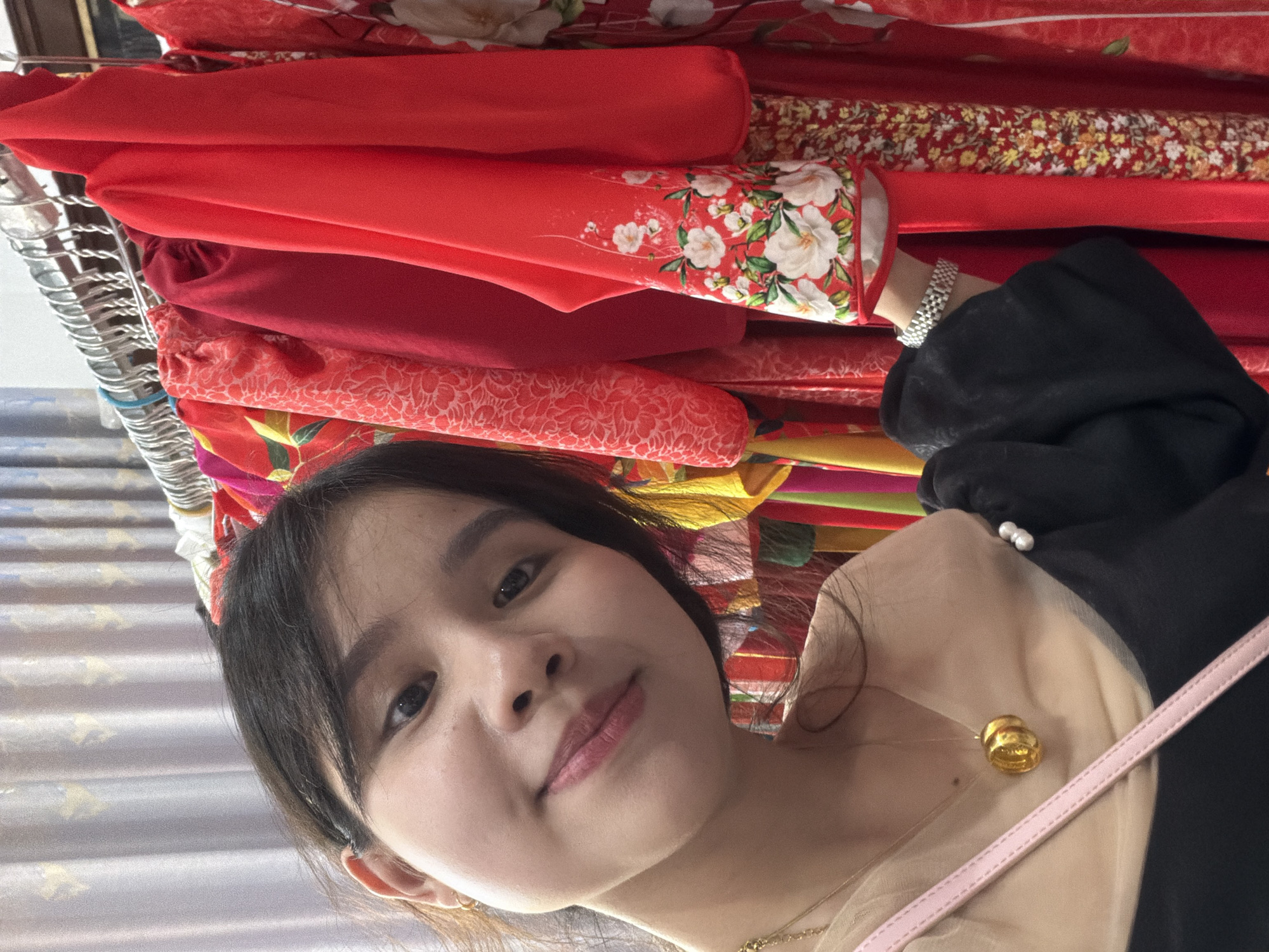 A young woman is featured in a slightly tilted selfie, looking directly at the camera with a pleasant, soft smile. Her dark hair frames her face, and she appears to be wearing a light-colored, sheer top layered under a black, long-sleeved garment. A delicate gold-toned necklace with a small, round pendant hangs around her neck, and a pink strap, likely from a bag, is visible over her right shoulder. On her right wrist, a silver-toned, multi-link watch or bracelet is visible as her hand reaches into a dense display of fabrics behind her.
The setting appears to be an indoor market or shop, likely in Bình An, Vietnam, filled with racks of textiles. The background is dominated by a vibrant array of fabrics and clothing, primarily in various shades of red. These materials showcase diverse patterns: one prominent fabric features large white floral designs with green leaves on a bright red background, while another has smaller, densely packed floral patterns in yellow, red, and white. There is also a solid red fabric and one with a subtle, lighter red embossed-like design. A metal display rack is partially visible on the far left. The overall scene suggests a casual shopping or browsing experience, with the woman appearing content. No discernible text is visible other than faint markings on the gold pendant.