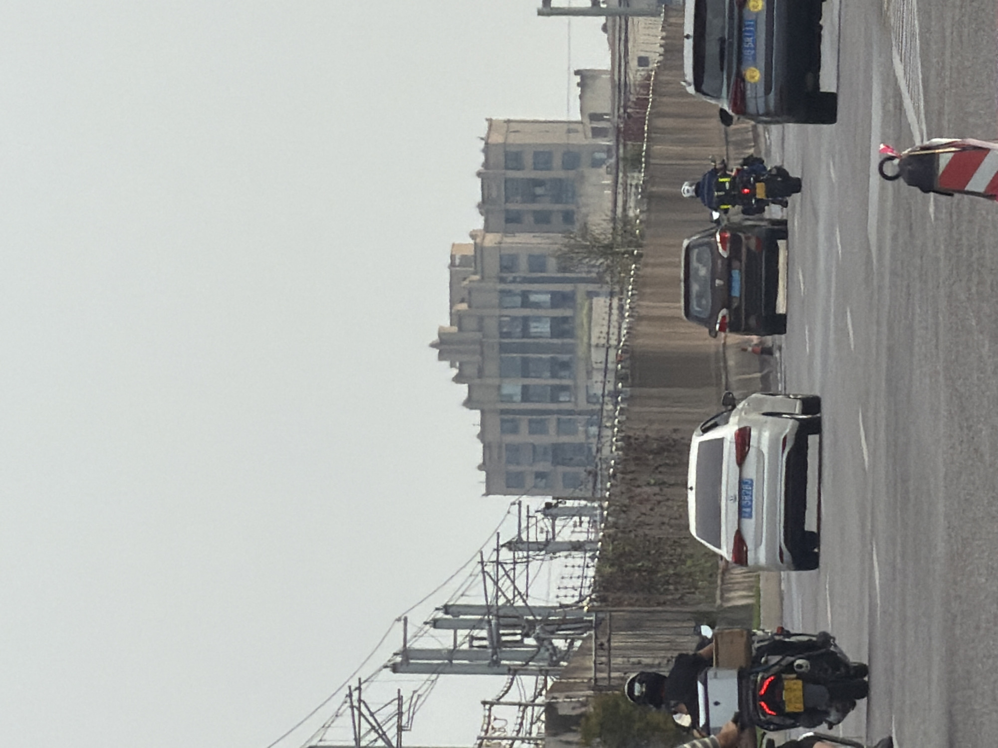 The image shows a view of a city street with traffic. In the foreground, there are several vehicles, including a white sedan, a dark-colored SUV, and two motorcycles. One of the motorcycles has a rider wearing a black helmet and a white box attached to the back. The other motorcycle has a rider with a white helmet and yellow luggage. In the background, there is a large, multi-story building with a distinct architectural style, possibly an old factory or apartment complex. Power lines and supporting structures are visible on the left side of the image. The road appears to be a multi-lane highway with white lane markings. The sky is overcast and appears hazy. This scene is consistent with traffic on a road in the city of Jiangjin, China.
