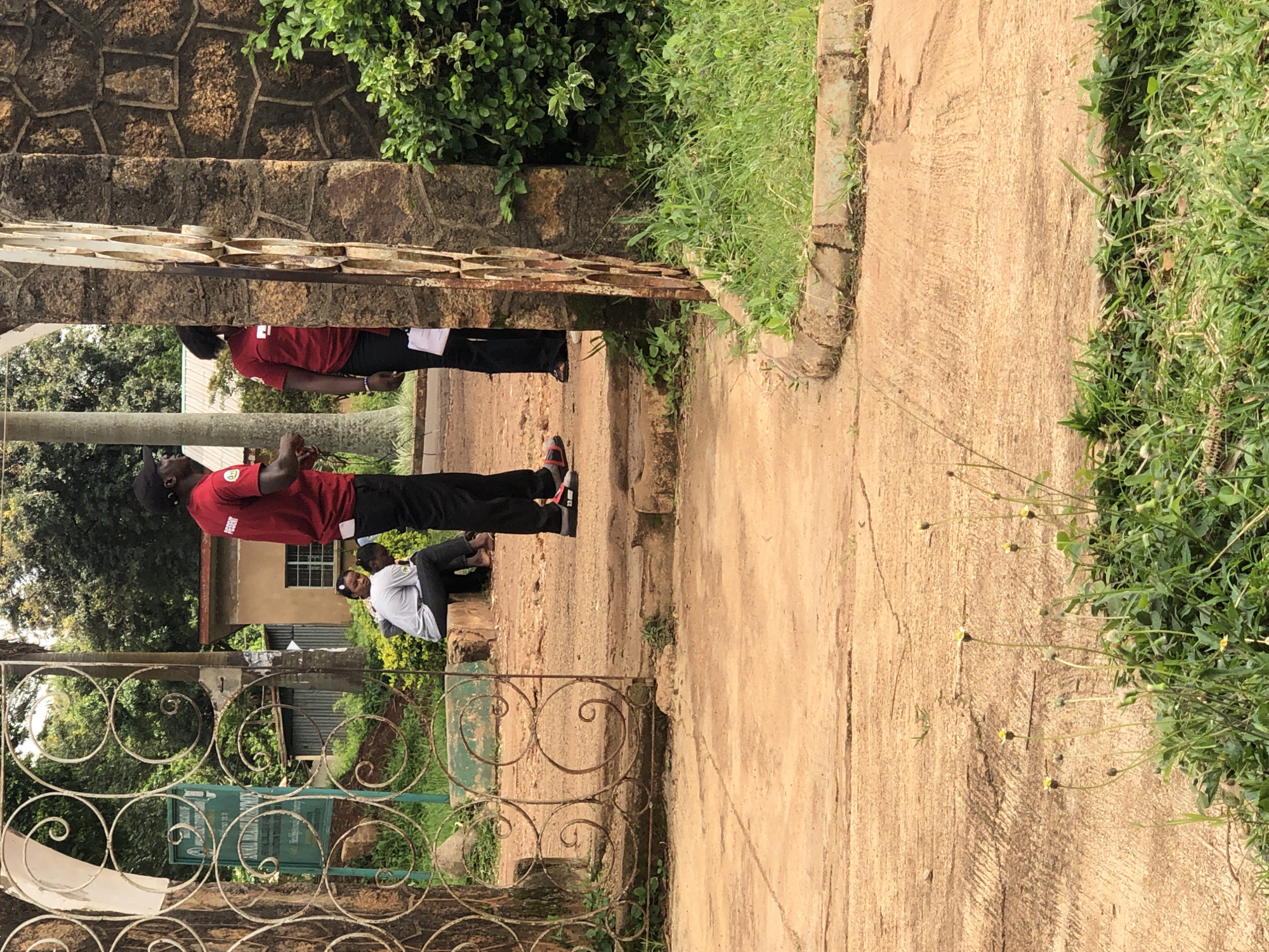 The image shows an outdoor scene in Vom, Nigeria, likely near a park or public area with a decorative metal gate. In the foreground, there is a concrete path with some cracks and patches of green grass and small yellow flowers. To the right, a stone pillar stands next to lush green bushes.
In the background, through the gate, two individuals wearing red shirts and black pants are visible. One is facing away from the camera, appearing to be speaking to the other person, who is partially obscured by the gate and a stone structure. They both seem to be standing. Further inside the gate, a third person, possibly a young person, is seated on a low wall or ledge, wearing a grey shirt and dark pants. Behind them, a small building with a window is visible, along with some trees and other vegetation. The overall impression is of a casual outdoor gathering or a moment of interaction in a public space.
