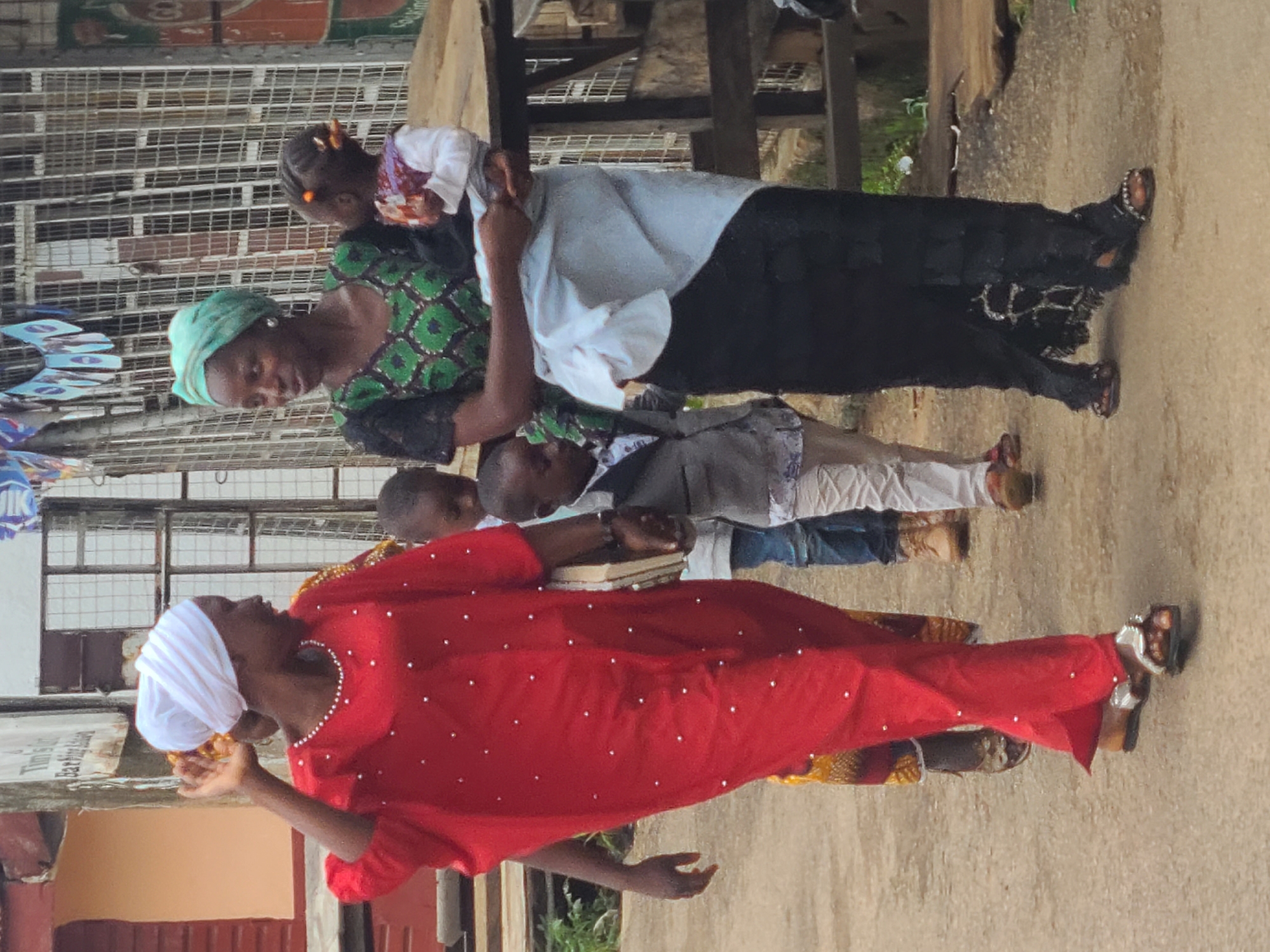 In the city of Jos, Nigeria, an outdoor scene captures a group of individuals walking along a dirt path. The focal point is a woman in a vibrant red, beaded kaftan, her white headwrap framing her face as she gestures with her left hand. Beside her, another woman, wearing a green and black patterned top and a turquoise headwrap, holds a baby. Two young boys, one in a grey suit and the other in a blue shirt and jeans, walk alongside the women. In the background, a metal fence and a wooden structure suggest an urban or semi-urban setting. The ground is a dusty, light brown, indicating dry conditions. The lighting is even, suggesting an overcast day or a shaded area.