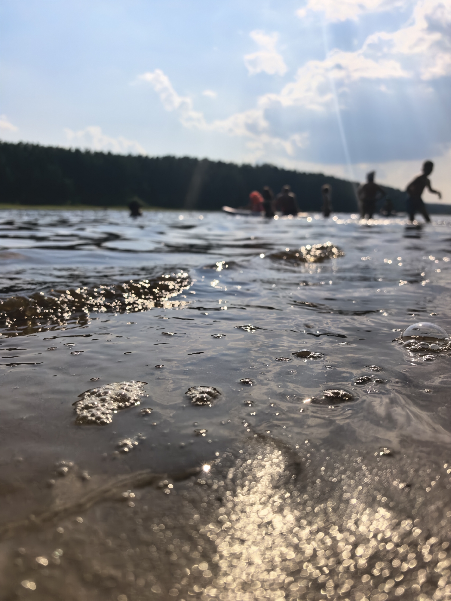This media file depicts a group of people enjoying a sunny day at a lake, likely during the summer. The camera is positioned low to the water, offering a close-up view of the rippling surface with sunlight glinting off the waves and creating bokeh effects. In the background, silhouetted figures are visible in the water, some appearing to be children. The shoreline is lined with a dense, dark green forest, suggesting a natural, undeveloped setting. The sky is blue with scattered, fluffy white clouds, and a bright sun casts rays through the atmosphere. The overall mood is one of leisure and outdoor recreation. The time of day appears to be afternoon, given the angle of the sunlight and the bright, clear weather. The location context of Sysert, Russia, points to a potentially rural or forested area known for its lakes. There is no visible text in the image.