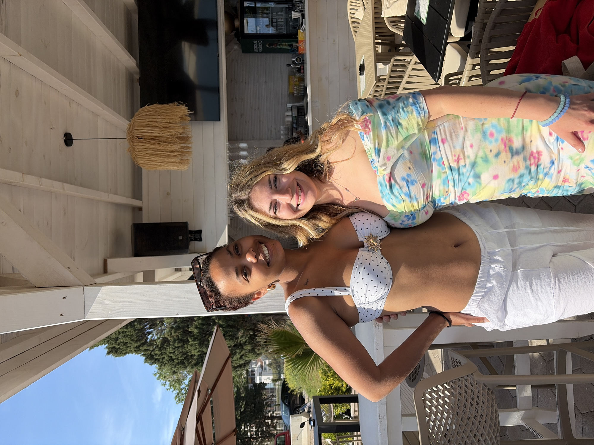 This portrait-oriented image, captured in Budva, Montenegro, features two women posing happily outdoors under a bright, sunny sky. The original image is rotated 90 degrees clockwise.
In the foreground, a woman with dark, curly hair, adorned with sunglasses on her head, smiles broadly at the viewer. She is wearing a white bikini top with black polka dots, featuring a distinctive golden sun-shaped embellishment at the center, paired with flowy white wide-leg pants. Her right hand rests on her hip, conveying a relaxed posture.
Standing slightly behind and to her right, a second woman with long blonde hair also smiles warmly. She is dressed in a light-colored, tie-dye patterned dress with floral motifs in shades of blue, yellow, and pink. On her left wrist, she wears a light blue beaded bracelet and a thin red string bracelet. A delicate necklace with a small pendant is visible around her neck.
The setting is an open-air establishment, likely a bar or cafe, characterized by a white wooden beamed structure and white plank walls. From the ceiling hangs a unique pendant lamp with a natural, straw-like shade. In the background, a bar area is visible with bottles, glasses, and a dark commercial refrigerator displaying text in Cyrillic, possibly "НЕПОВ Е НДКО КИЧКО". A large black flat-screen television is mounted nearby. Beige outdoor chairs and tables are present throughout the scene. Beyond the structure, vibrant green trees, a clear blue sky, and other outdoor resort elements like a white canopy and a vehicle are glimpsed. Both women exude joy and relaxation, suggesting a pleasant vacation or leisure moment. The strong sunlight indicates a daytime scene with clear weather conditions.