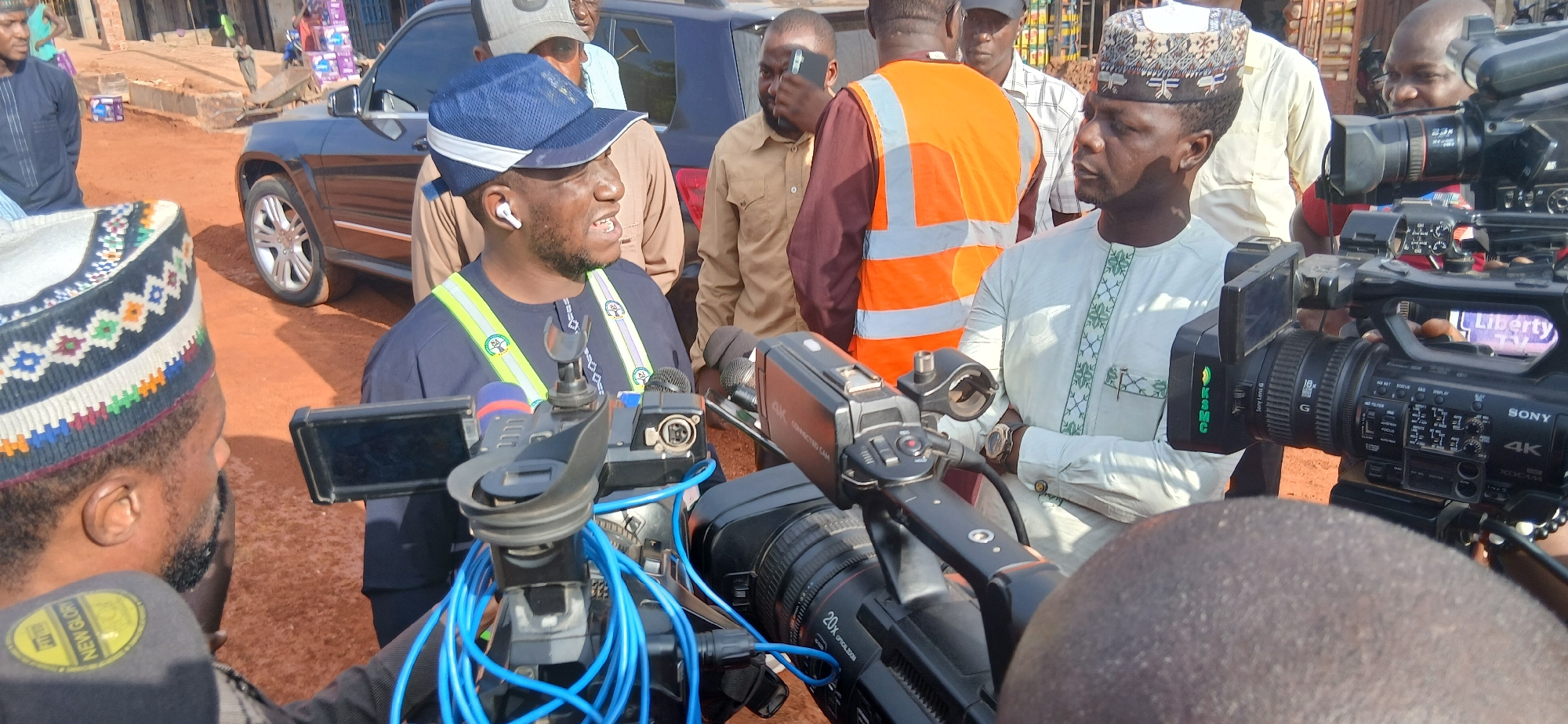 In Zaria, Nigeria, a group of men are gathered outdoors on a dirt road, suggesting a news reporting or public relations event. The central figure, wearing a blue cap and a dark blue shirt with reflective yellow and green stripes, is speaking. He is wearing white earbuds and has a microphone attached to his shirt. In front of him are several professional video cameras, some with "SONY 4K" branding, indicating a media presence. Other individuals in the scene include a man in a light-colored kaftan with embroidered patterns, another in an orange safety vest, and others partially visible in the background, some holding phones or cameras. A dark-colored SUV is parked behind the central figure. The environment appears to be a dusty, unpaved urban area with some visible construction materials and stacked items in the background.
