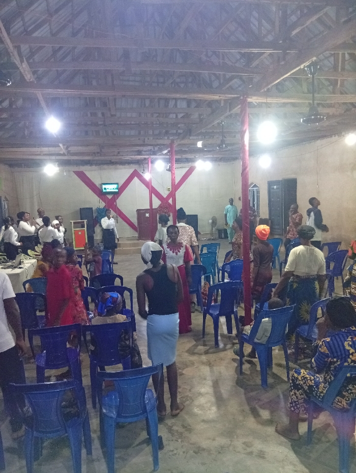 A group of people are gathered in what appears to be a church or meeting hall. Many are seated in blue plastic chairs, while others stand and talk. The room has a high, exposed wooden beam ceiling with several hanging lights and fans. Decorations include red fabric draped behind a stage or platform area, and a television screen is visible. The atmosphere seems relaxed and social.