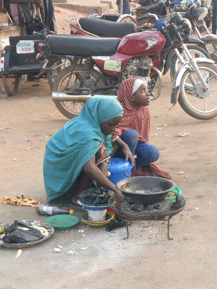 This image, captured in Alizaga, Nigeria, depicts two young females squatting on an unpaved, dusty ground, likely engaged in street vending activities. In the foreground, a female wearing a teal hijab and patterned top is actively tending to a dark, wok-like pan resting on a small, metal charcoal stove or grill. Ash is visible around the stove, indicating its use. To her left, several items are arranged on the ground: a black substance in a white bowl atop a yellow plate, a green plastic plate with liquid, an empty aluminum plate with white remnants, a dark bottle, and a folded piece of patterned fabric. A blue plastic bucket with a lid is positioned between the two individuals. The second female, to the right and slightly behind, wears a red and white patterned hijab and blue pants, looking upwards and to the right, observing the surroundings. In the mid-ground, multiple motorcycles are parked, including a prominent red motorcycle with a black seat and a sidecar-like attachment displaying "WOFAN ICON". Other blurred figures are visible in the background. The overall environment is an outdoor, earthy setting typical of a marketplace or street.
