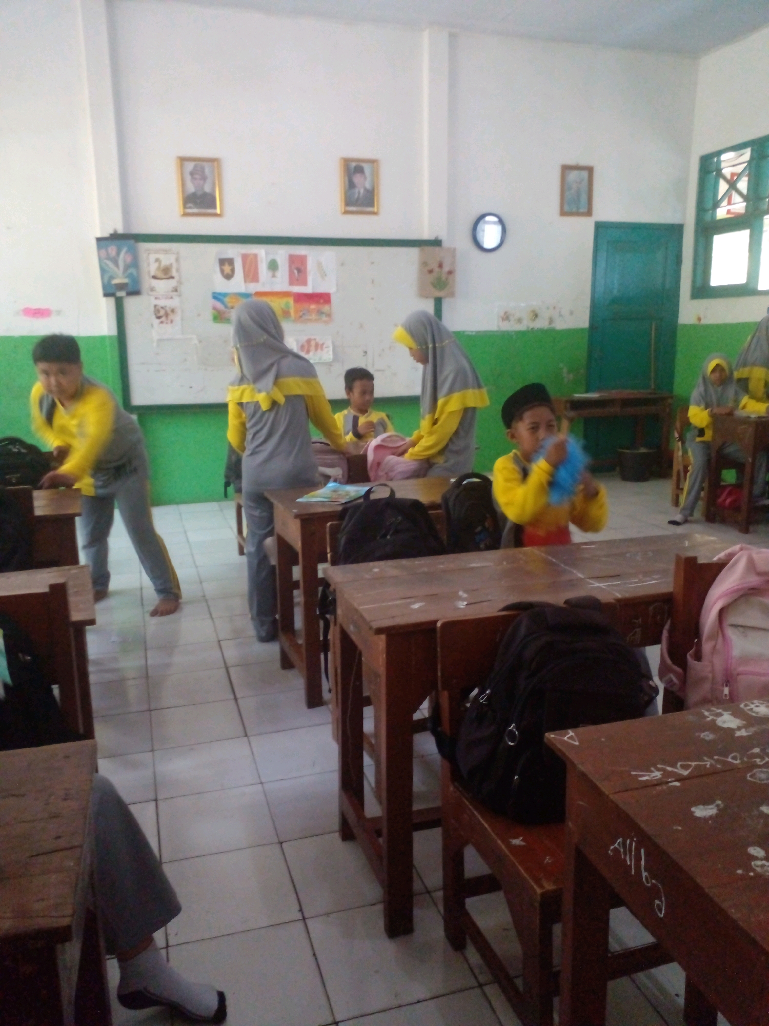 The image depicts an indoor scene, likely a classroom, in Banyuputih, Indonesia. The room is sparsely decorated, with white walls and green wainscoting. Several wooden desks and chairs are arranged in rows, and some students are present.
In the foreground, a student wearing a yellow shirt and grey pants is partially visible, with a grey sock and black sandal on their left foot. To the right of this student, another student in a yellow and grey uniform is seated at a desk, holding a blue fluffy object. In the background, a student in a yellow shirt and grey pants is walking away, and another student in a similar uniform is visible near a desk.
The walls of the classroom are adorned with framed pictures and drawings. One framed picture shows a person in traditional attire, and another appears to be a portrait of a historical figure. There are also several unframed drawings and papers displayed. A blue circular clock is mounted on the wall.
A green wooden door is visible in the background, leading to an area that appears to be outside. A window with green shutters is also present on the right side of the room. The floor is tiled with light-colored square tiles. The overall impression is of a simple and functional educational environment.