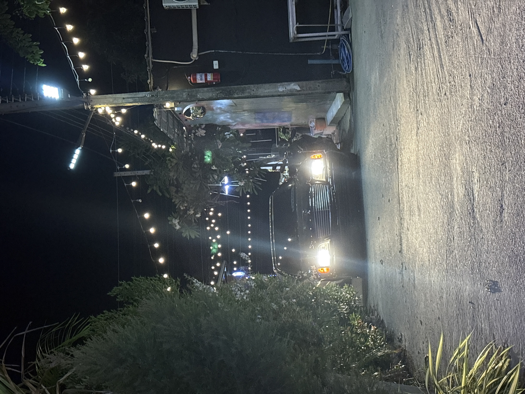 Baan Bang Rak, Thailand. Nighttime. A dark-colored vehicle, likely a passenger van or bus, is parked next to a building with a textured gray wall. The vehicle's headlights are on. A string of lights is draped overhead and along a path leading away from the vehicle. A fire extinguisher is mounted on the wall above the vehicle. Trees and shrubs are in the foreground.