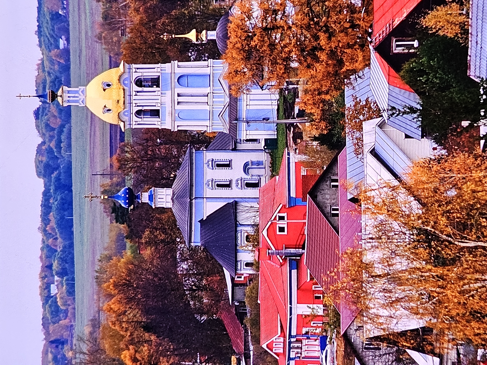 This is an aerial, medium-distance shot of a rural landscape in Vladimir, Russia, during autumn. In the foreground, a collection of red-roofed houses and buildings with various colored roofs are visible, interspersed with trees displaying vibrant orange and yellow foliage. Behind these structures, a large, white and blue Orthodox church with a golden dome stands prominently, accompanied by a smaller blue-domed church. The background consists of rolling hills covered with trees in autumn colors and fields of muted green and brown. The sky is a pale, overcast lavender color. No people or animals are visible in the frame.