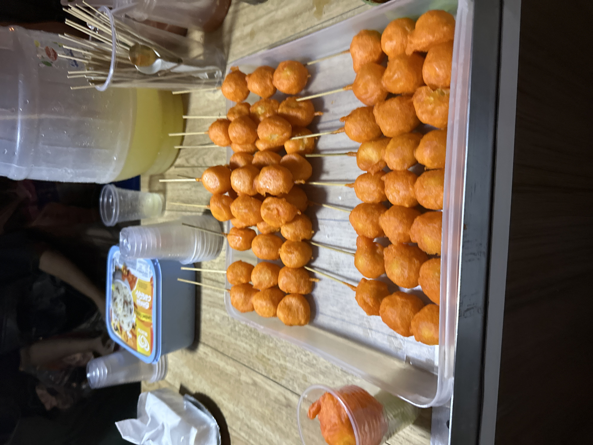 The image captures a casual food setup, likely indoors, featuring numerous skewers of orange-colored, deep-fried food, commonly known as Kwek-kwek or similar Filipino street food, consistent with the Davao City, Philippines location.
The primary subjects are rows of small, round, orange-hued items, each threaded onto wooden skewers. These skewers are neatly arranged in a long, clear plastic serving tray, filling much of the right side of the frame. To the upper left, a large, translucent plastic dispenser contains a yellow liquid, likely a beverage such as lemonade or juice. A collection of thin wooden skewers is housed in a clear cup beside the dispenser, ready for use.
Other objects visible include several clear plastic drinking cups, some stacked, scattered across a light-colored wooden surface. Towards the lower left, a blue-lidded rectangular container is present, possibly holding a dessert or snack item. Visible text on this container's label includes "SHAKE CRAVE" and "Limited Edition." A white tissue or napkin dispenser is also partially visible on the bottom left. One of the orange skewered items is shown placed inside a clear plastic cup on the bottom right, suggesting individual serving.
The scene depicts a food preparation or serving area for a gathering or event. The overall impression is that of an informal setting where these popular snacks are being offered for consumption. The lighting suggests an indoor environment, possibly in the evening, with artificial light illuminating the setup. No specific individuals or strong emotions are discernible, but the arrangement implies social interaction around food.