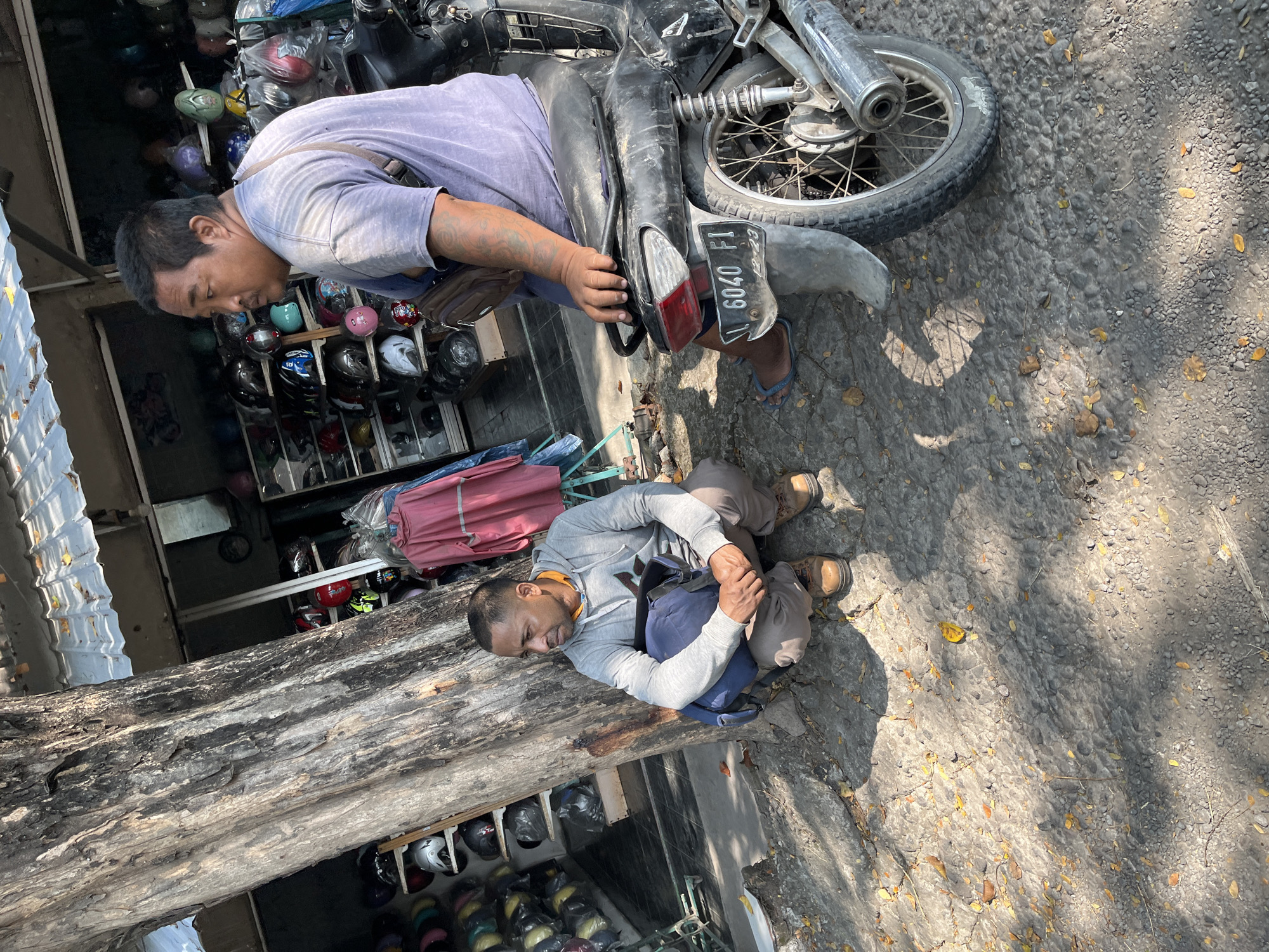 A man is sitting on a motorcycle, looking away from the camera. Another man is squatting next to a tree, with a backpack at his feet. The scene appears to be in front of a shop selling helmets, with shelves of helmets visible in the background. The ground is rough, a mix of gravel and stone, with scattered leaves. The overall environment suggests a street-side setting in a warm, possibly tropical climate, consistent with Kludan, Indonesia. The lighting indicates it is daytime, with dappled sunlight and shadows.