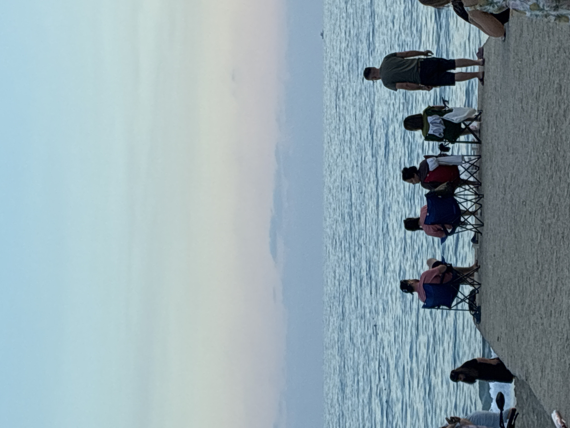 The image shows a group of people sitting in chairs on a concrete walkway next to a body of water. The water is calm with gentle ripples, reflecting the pale blue and white of the sky, suggesting it's either early morning or late afternoon. The sky is a gradient of soft blues and whites, indicating a clear and mild day, possibly with some light cloud cover.
The people are spaced out along the walkway, with several individuals sitting in folding chairs facing the water. One person, a man in a green shirt and dark shorts, stands on the walkway, looking out at the water. Further down, a woman is partially visible sitting in a folding chair. The clothing worn by the people suggests a casual, warm-weather setting.
The overall atmosphere appears relaxed and peaceful, with people enjoying the view of the water. The concrete walkway runs along the edge of the water, providing a vantage point for the onlookers. There are no visible signs or text in the image. The scene is likely a public waterfront area, common in coastal towns. The presence of folding chairs suggests a leisurely activity, such as fishing, watching a sunset, or simply enjoying the scenery. The image is oriented vertically, with the water and sky dominating the left side and the people and walkway on the right.
