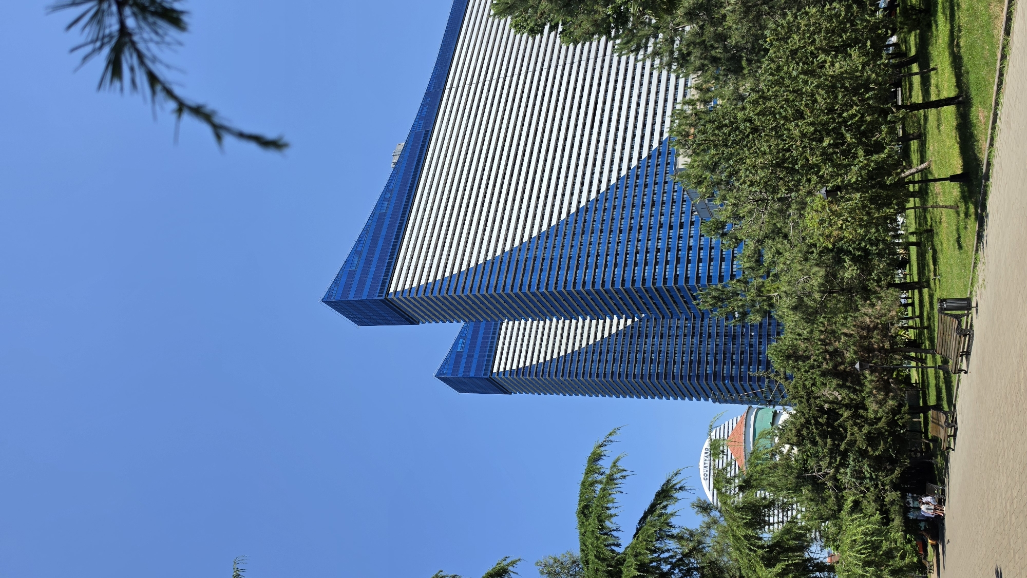 This is a daytime, eye-level, outdoor photograph of the Arch and Noah buildings in Batumi, Georgia. Two identical, modern, blue and white striped skyscrapers dominate the center of the frame, with a clear blue sky as the backdrop. In the foreground, a paved walkway leads through a lush green park filled with mature pine trees and benches. Two wooden benches are visible, one with a trash can next to it. A few people are seated on a bench in the distance, near a white building with the word "COURTYARD" visible on it. The scene appears peaceful and well-maintained.